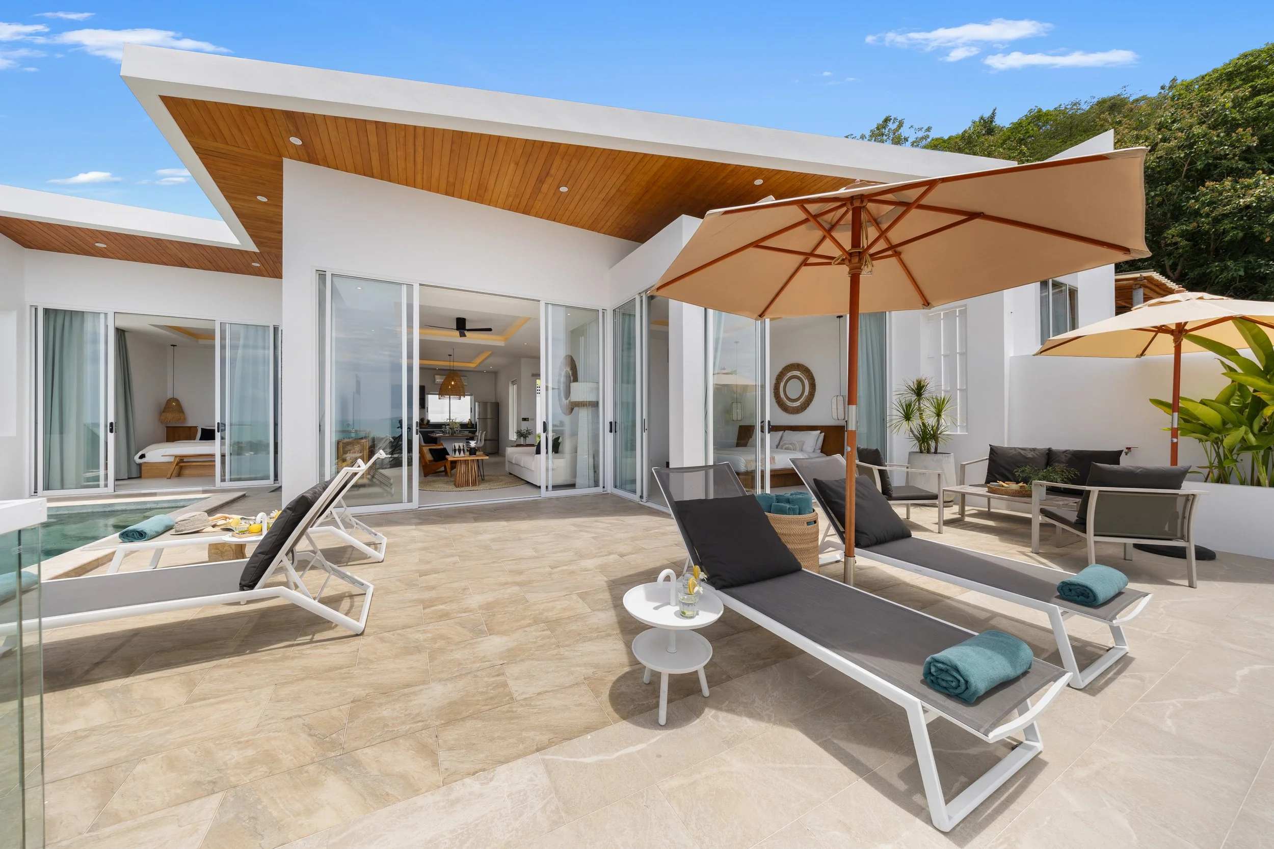 Modern house with white exterior, outdoor patio, lounge chairs with towels, umbrellas, and open sliding glass doors revealing a living room inside, trees in the background under a blue sky.