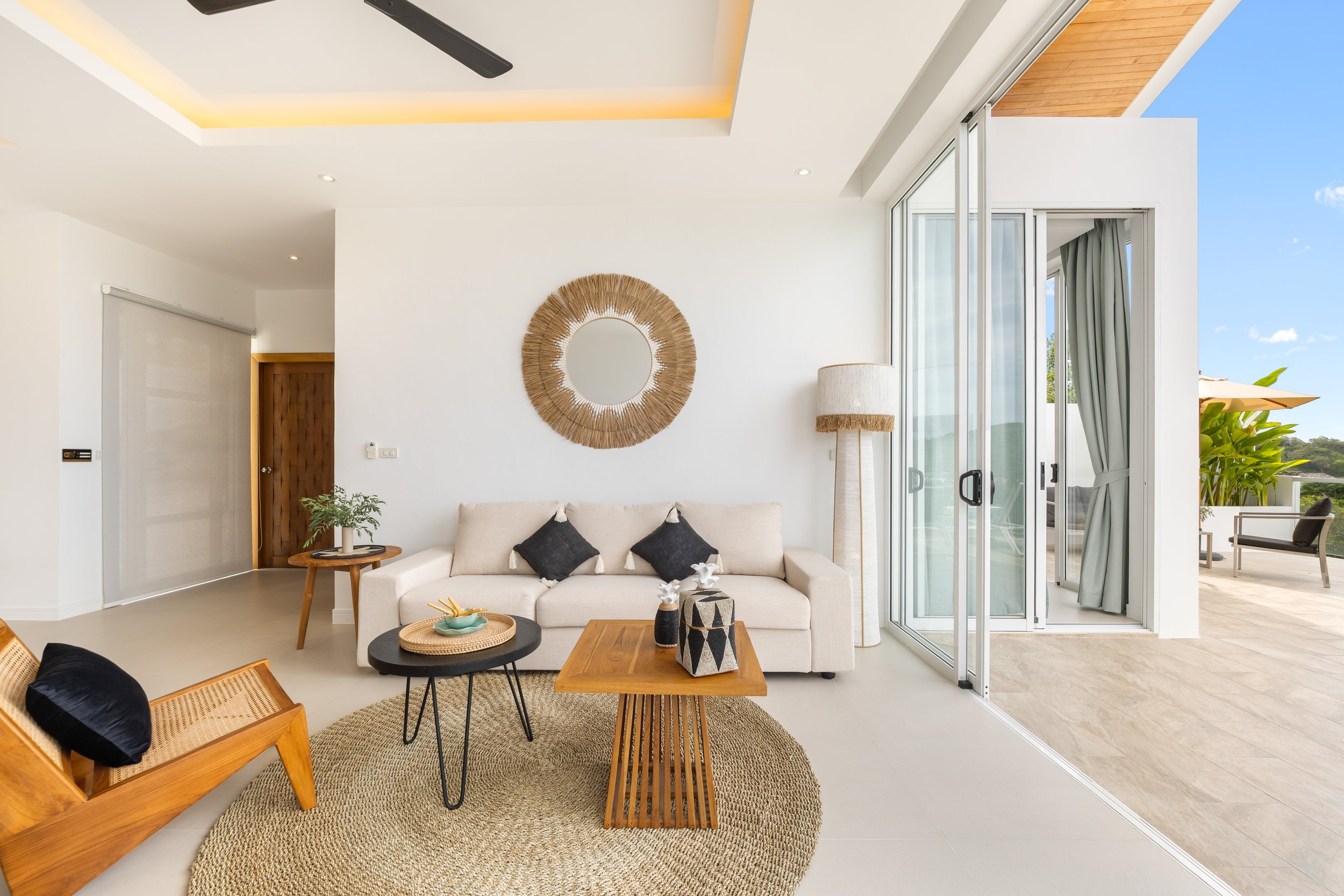 Living room with white walls, beige sofa with black pillows, round black coffee table with decorative vases, wooden side table with plants, large glass sliding doors leading to an outdoor patio, ceiling fan, and modern decor.