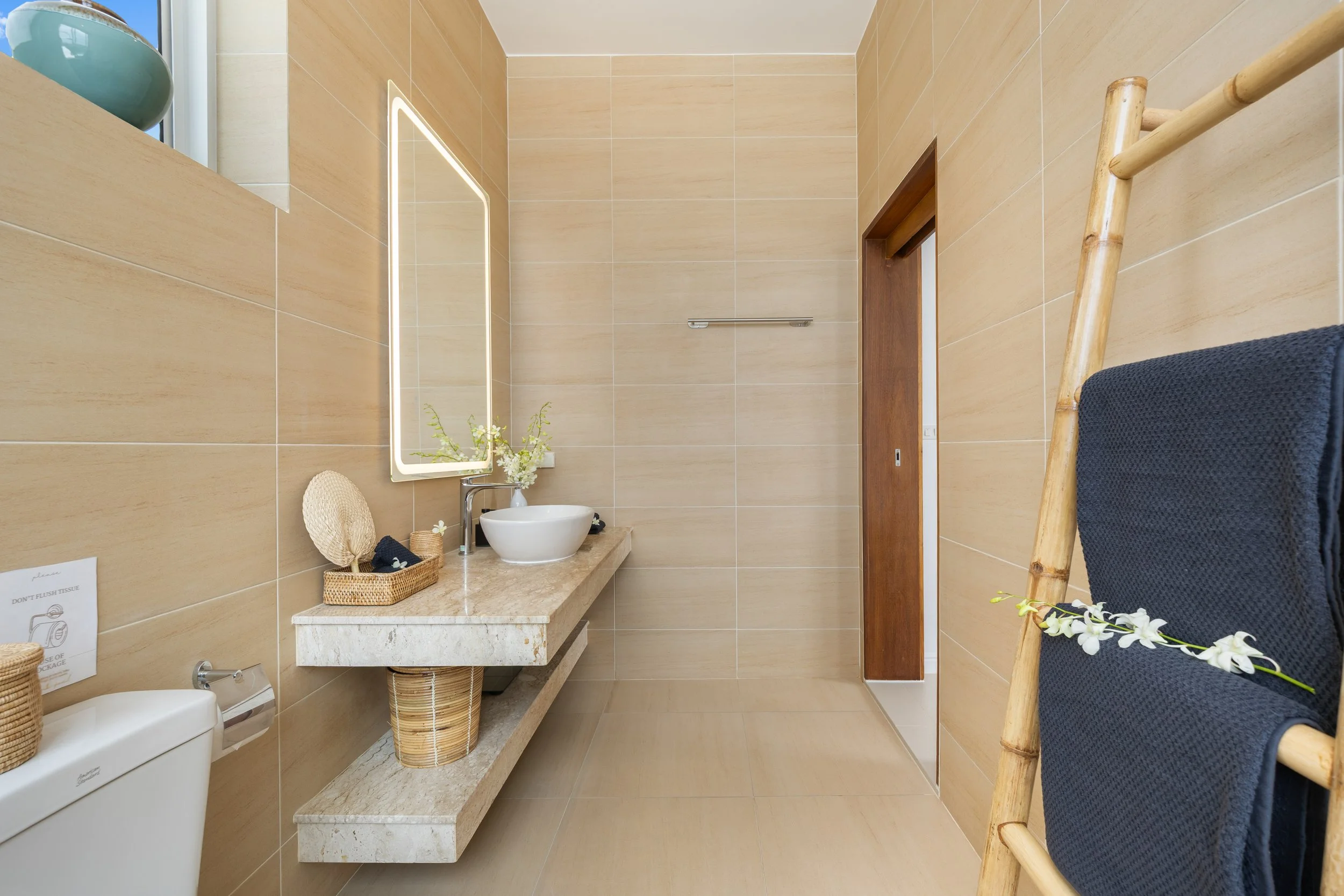 Bathroom with beige tiles, a mirror with lighting, a white vessel sink, a wooden door, a towel rack, a bamboo ladder with a navy towel, a flower arrangement, and a wicker basket.
