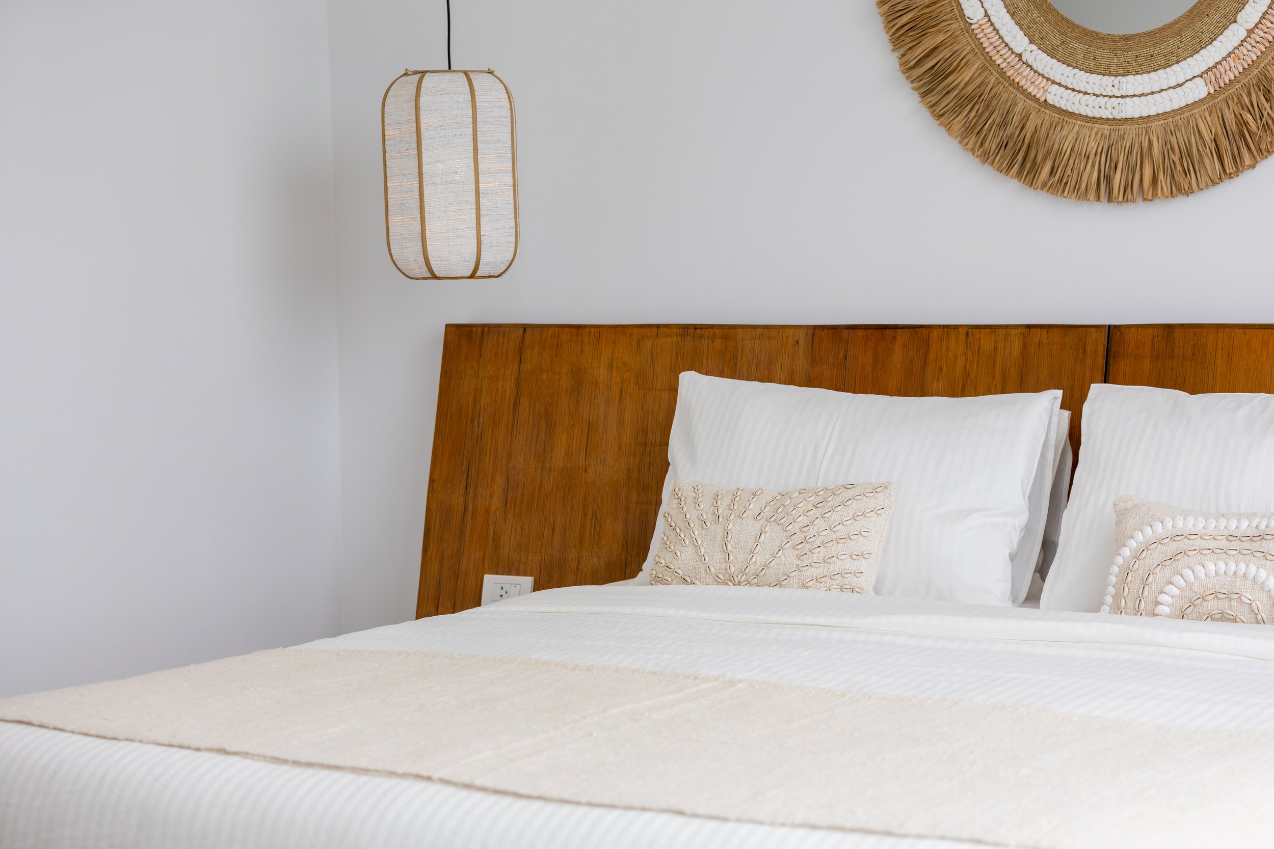 A neatly made bed with white linens, a beige decorative pillow, and a wooden headboard. A woven wall hanging and a hanging lantern are on the wall above the bed.