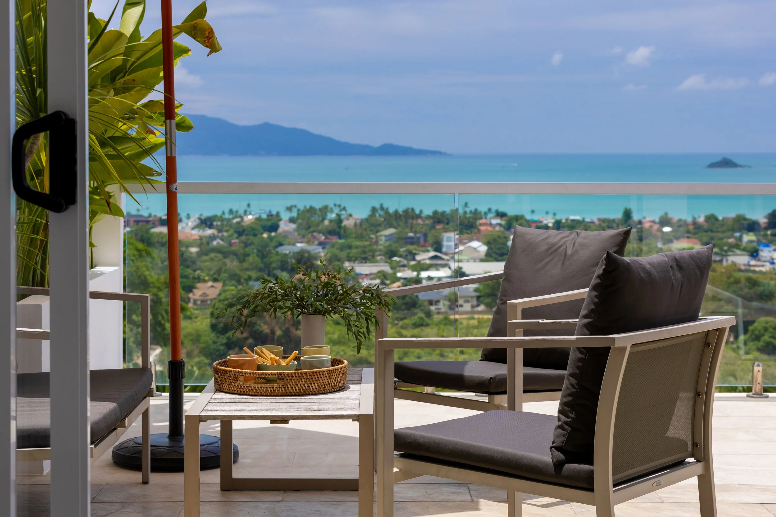 Balcony with modern chairs and pillows overlooking a coastal town, ocean, and distant mountains.