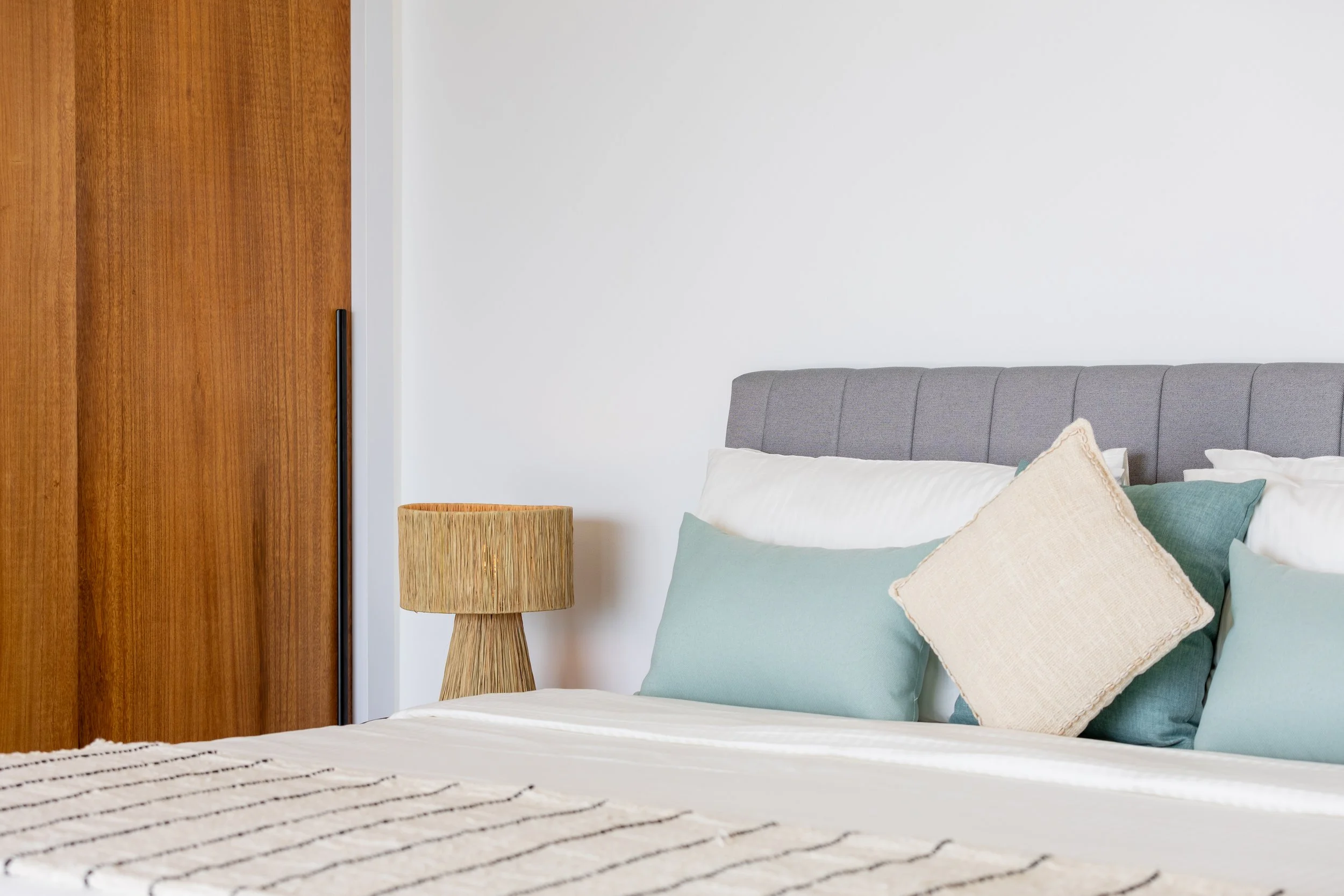 A bedroom corner with a wooden door, a bed with a grey upholstered headboard, multiple pillows in pastel colors, a wooden bedside table, and a woven table lamp against a white wall.