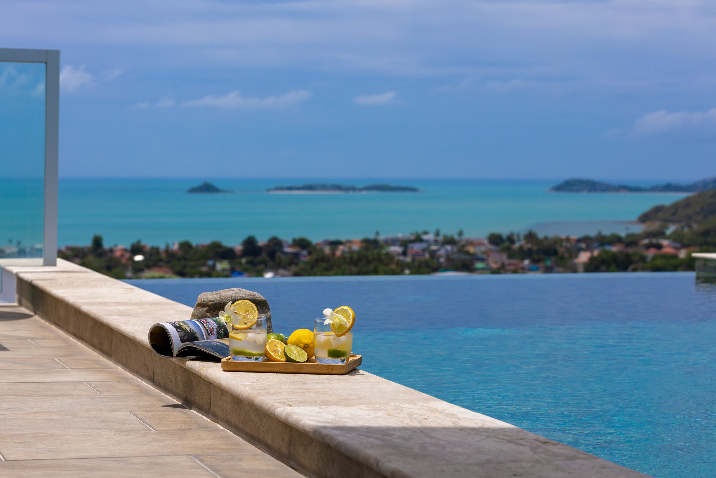 A tray with lemons and a lemon-flavored beverage sits on a stone ledge by a swimming pool with a view of the ocean and islands in the background.