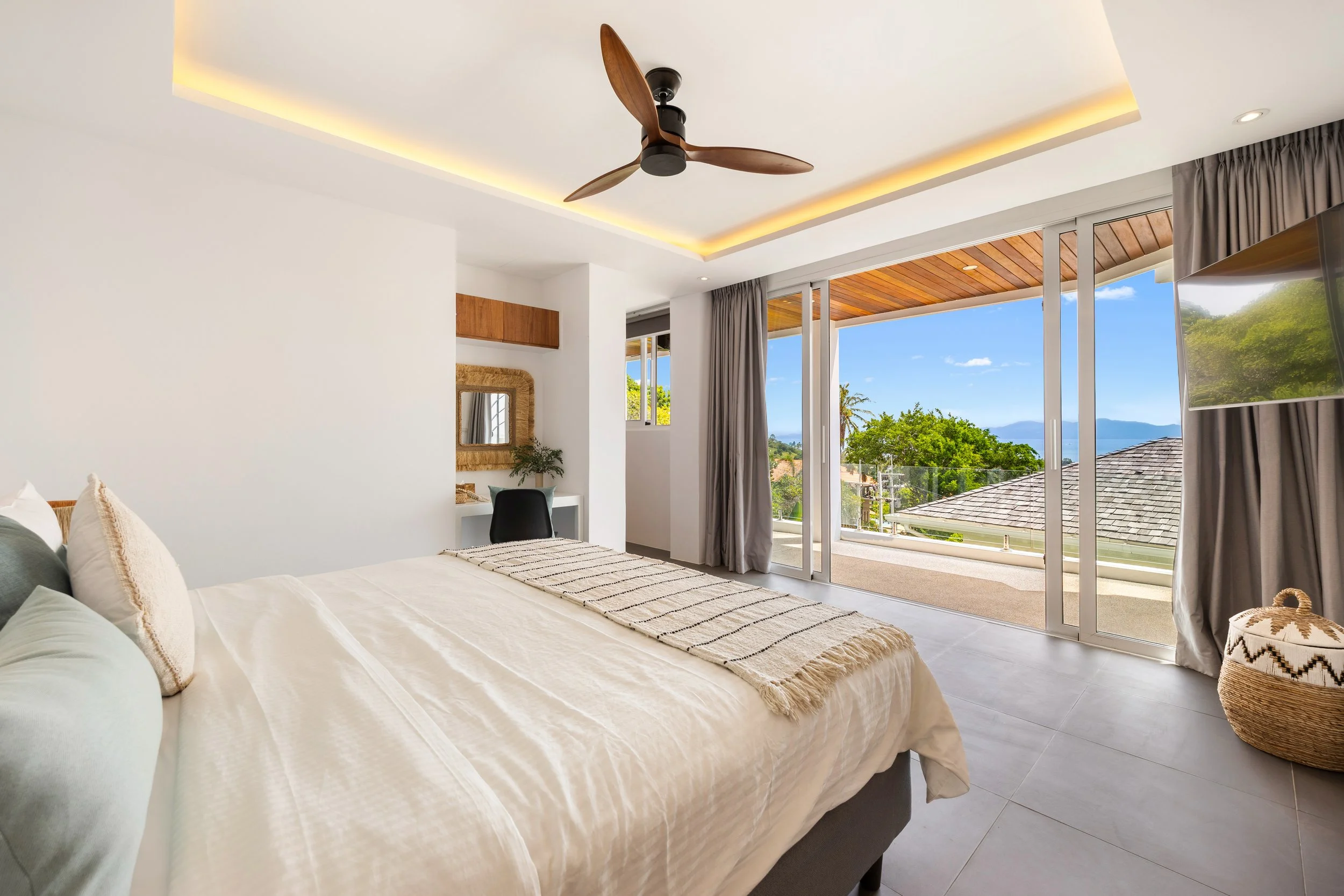 A bright bedroom with large glass sliding doors opening to a balcony, showing a scenic view of trees, roofs, and a body of water in the distance. The room has white walls, a ceiling fan, and light-colored furniture, including a bed with decorative pillows, a small desk, and a wicker basket.