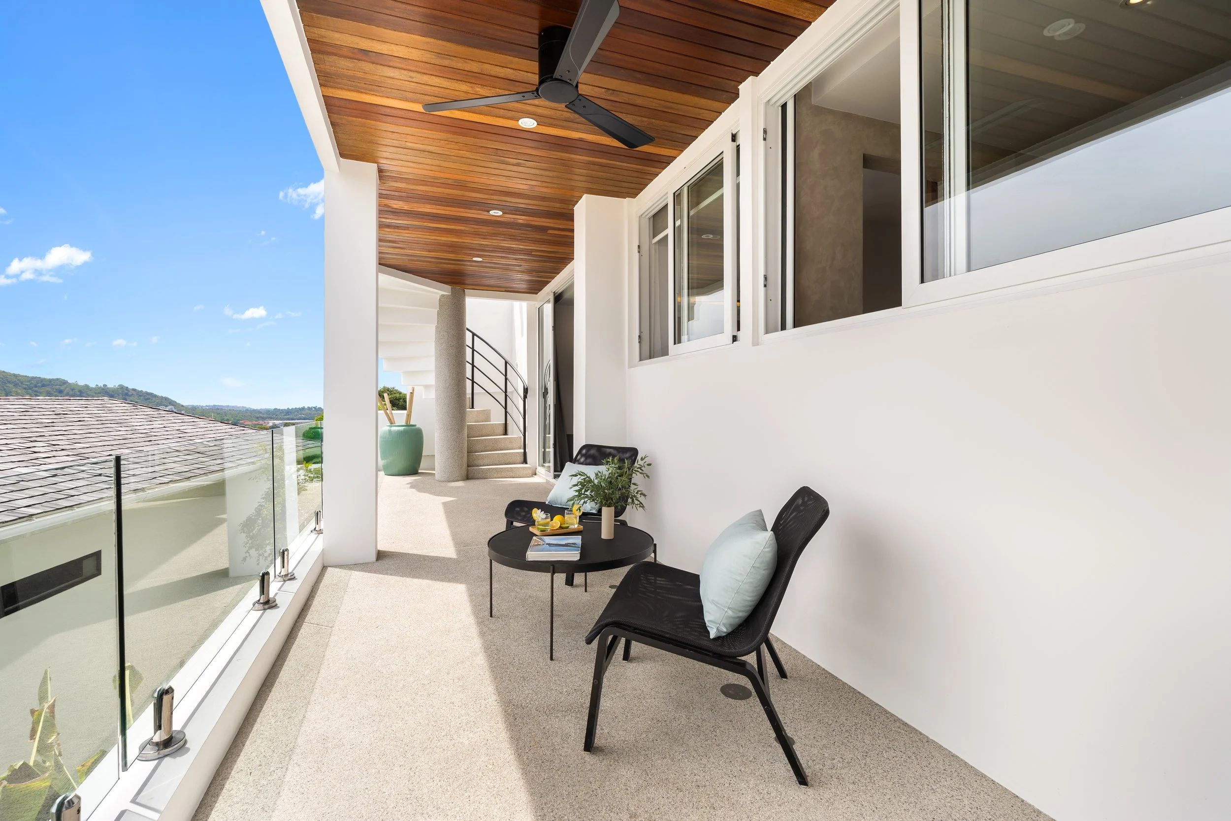 A modern balcony with beige carpet, black outdoor chairs with pillows, a small black round table with drinks and magazines, glass railing, and a white wall with windows and a wooden ceiling fan, overlooking a clear blue sky and distant hills.