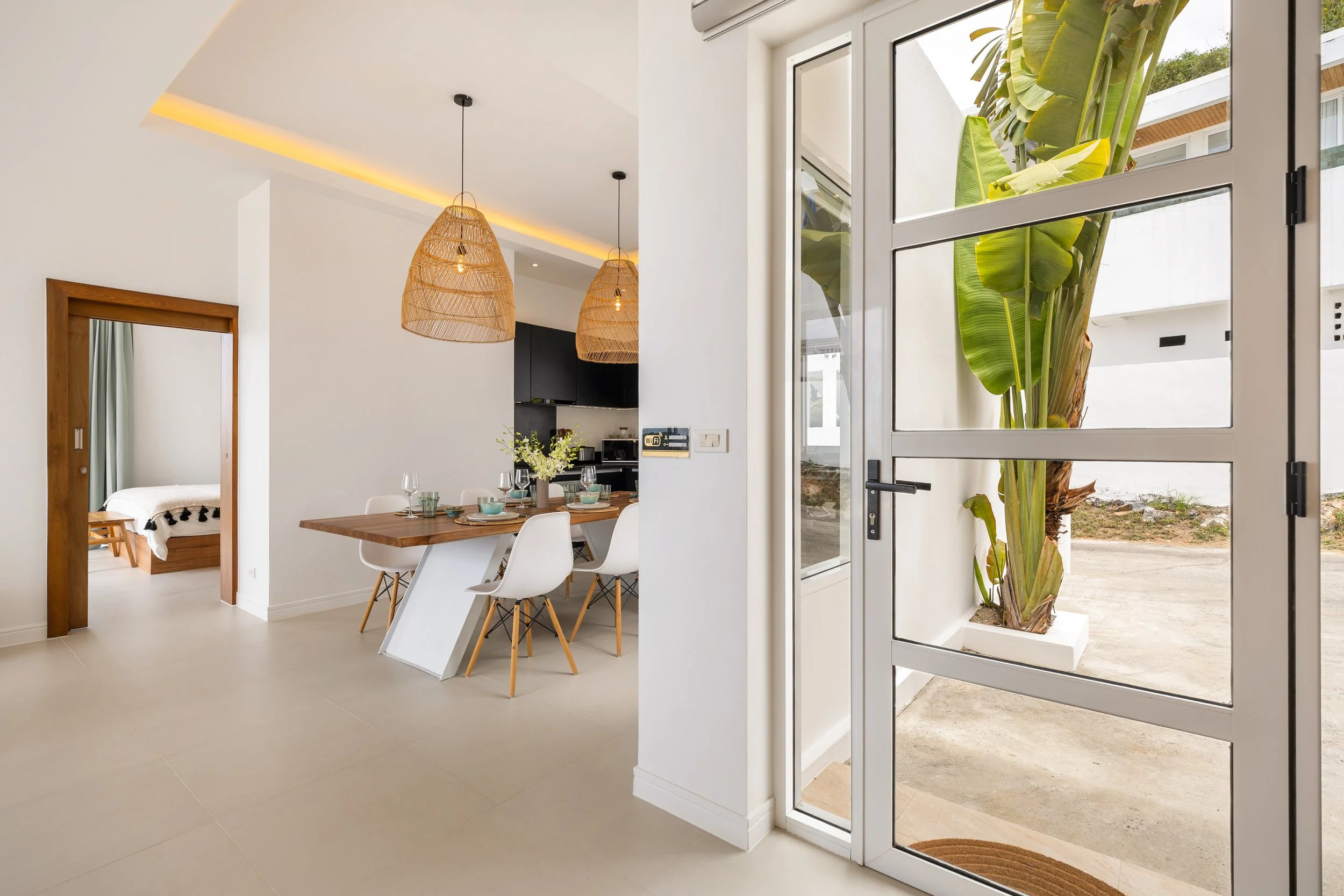 Modern interior with white walls, a wooden dining table set with plates and glasses, hanging wicker pendant lights, and large windows showing lush green plants outside.