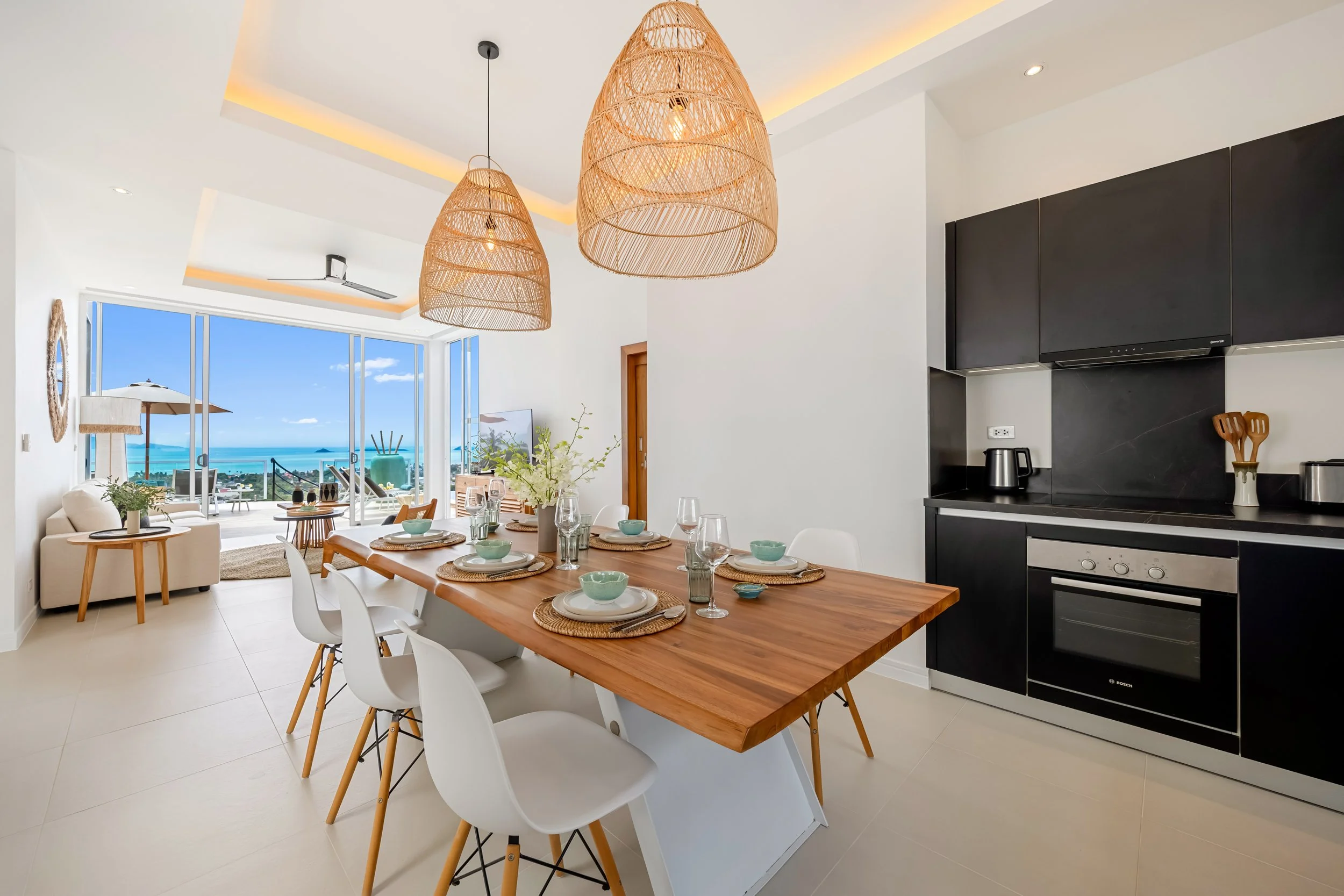 Bright open-concept living and dining area with large windows overlooking the ocean, featuring a wooden dining table set with dishes and glasses, white chairs, black kitchen cabinets, and woven pendant lights hanging from the ceiling.