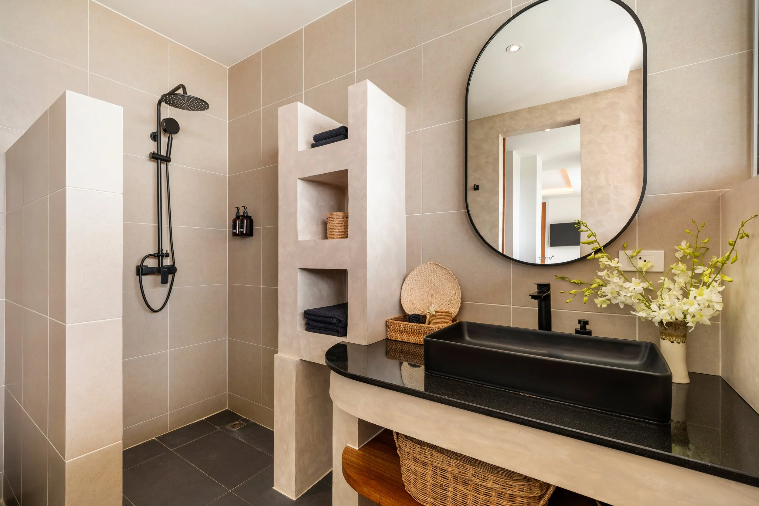 Modern bathroom with beige tiled walls, black sink, large oval mirror, built-in wall niches with towels and accessories, black shower fixtures, small wicker baskets, and a vase of white flowers.