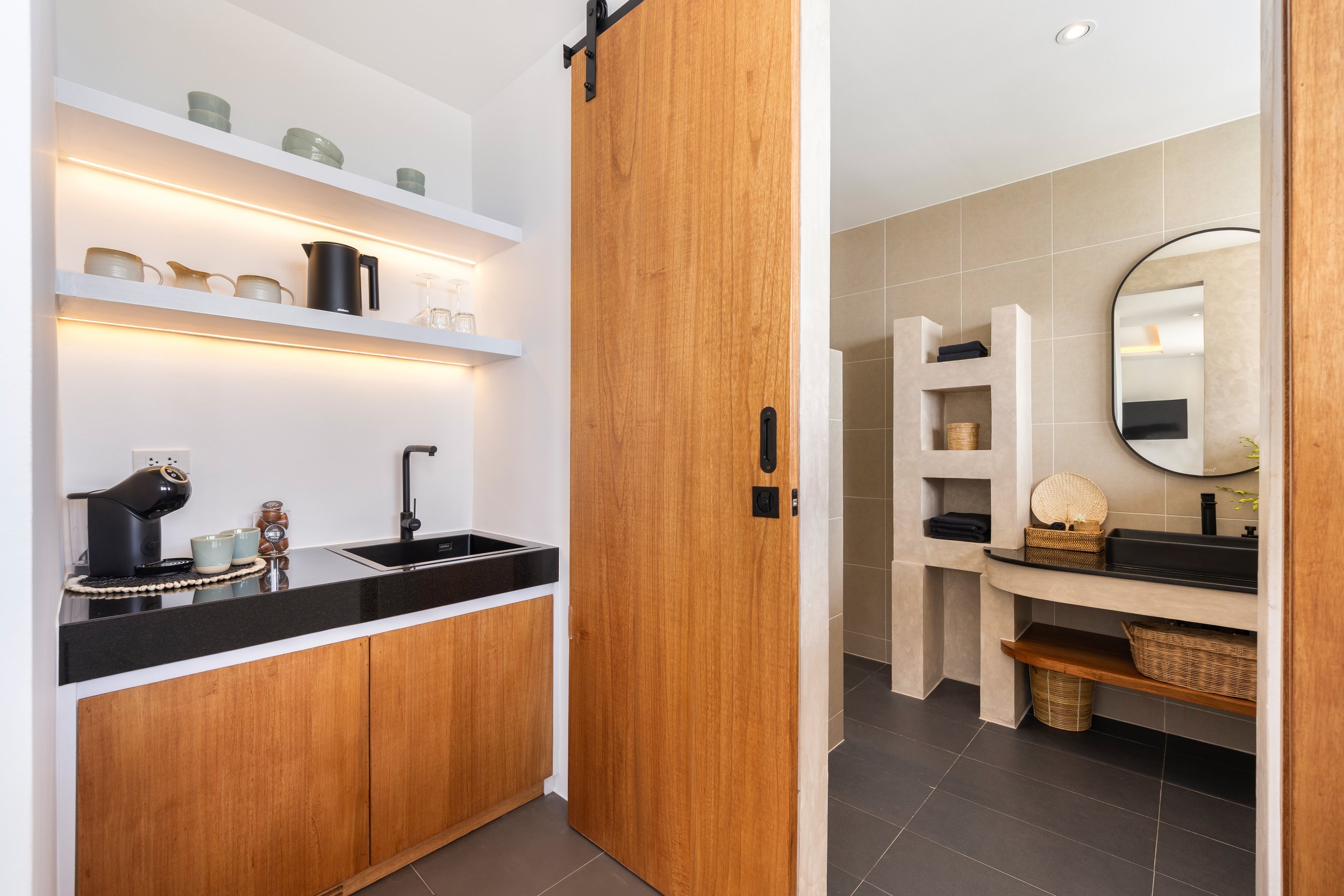 A modern kitchenette with wooden cabinets, a small sink, black countertop, and open shelves with dishes and glasses. Next to the kitchenette is a bathroom area with a round mirror, beige tiled walls, a built-in shelf with towels and decor, and a wicker basket.