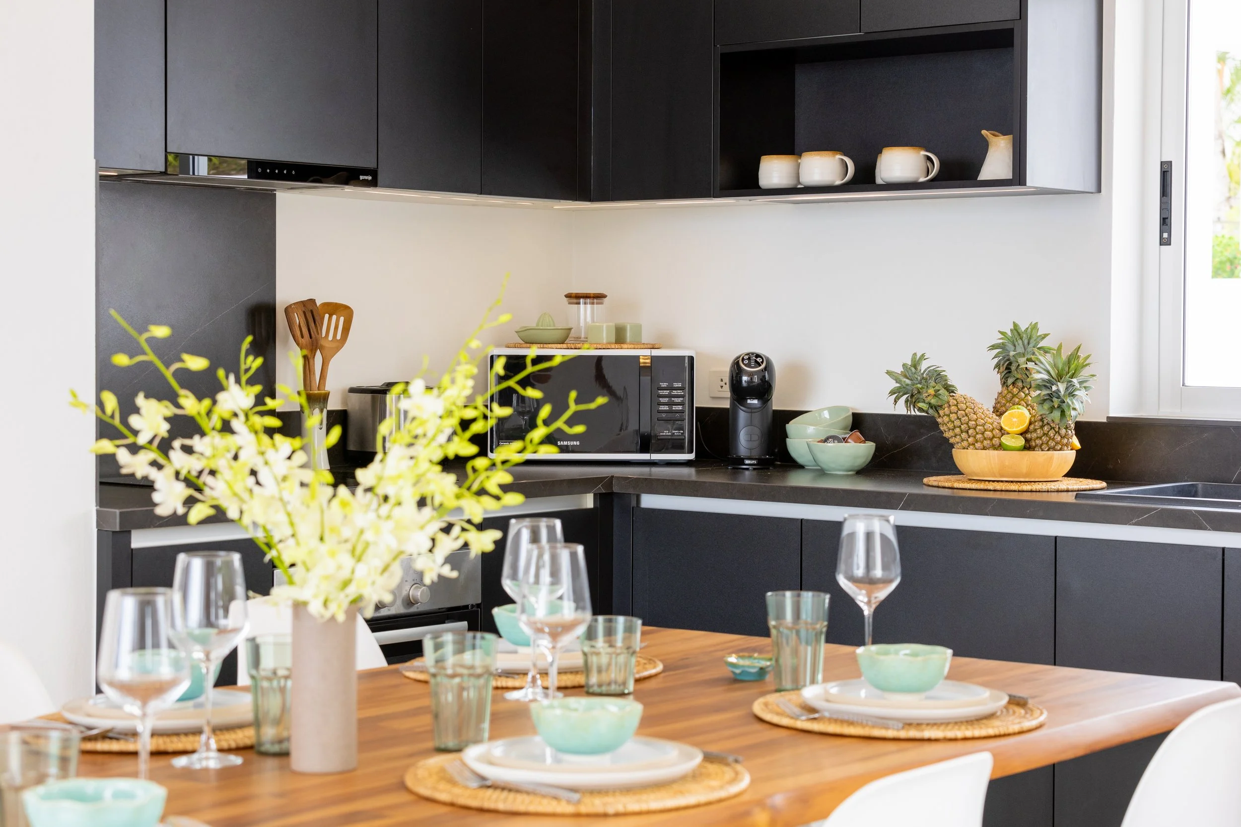 A modern kitchen and dining area with black cabinets, a wooden dining table set with glassware, small bowls, and a floral centerpiece. The kitchen counter has a microwave, a fruit bowl with pineapples, and a coffee machine. There are decorative cups and a pitcher on the upper cabinets, and a window on the right letting in natural light.