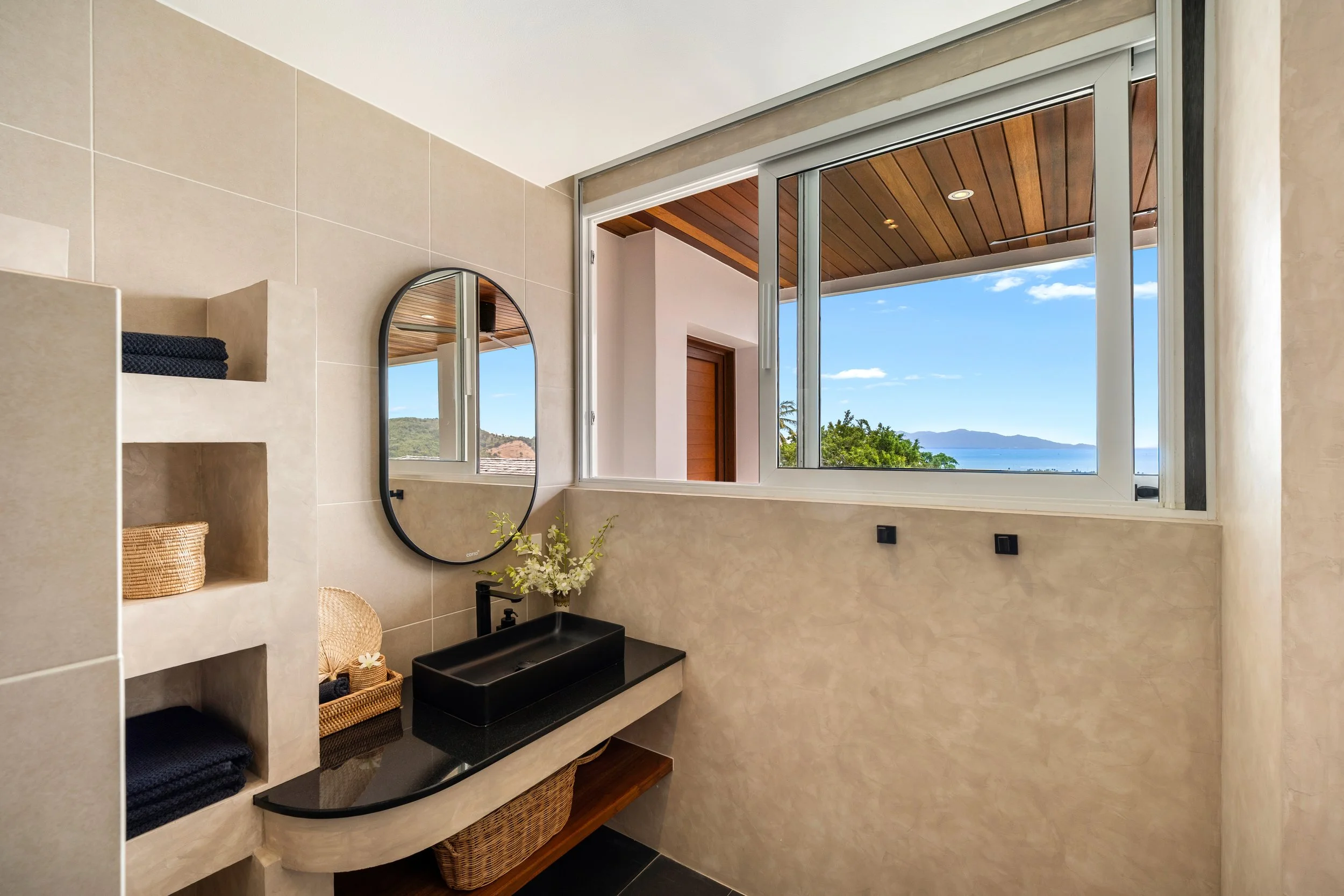 A modern bathroom with large windows showing a scenic view of blue sky, mountains, and water, beige tiled walls, a black sink, and woven baskets.