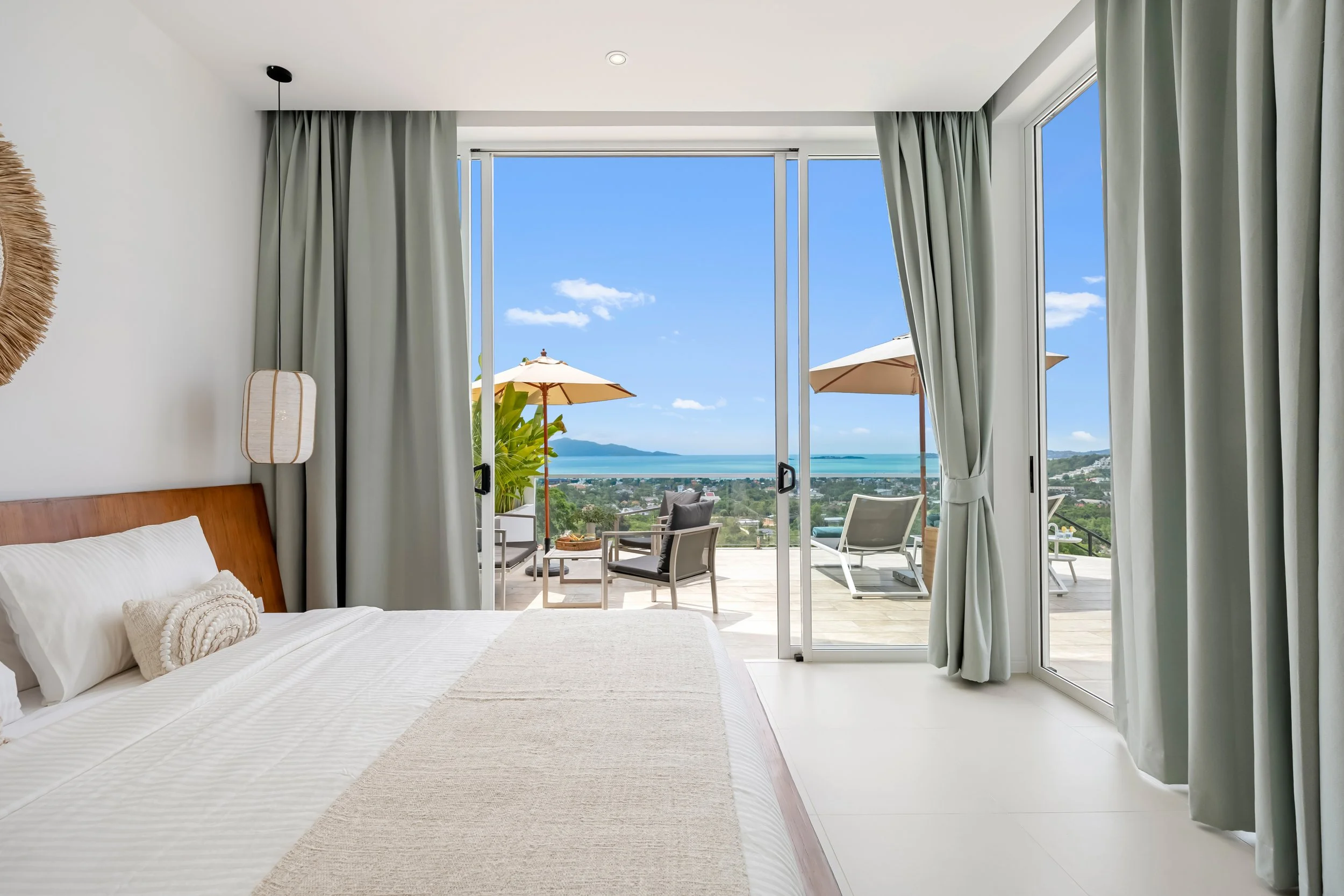 Bedroom with large glass sliding doors opening to a balcony with outdoor furniture and umbrellas, overlooking a scenic ocean view.