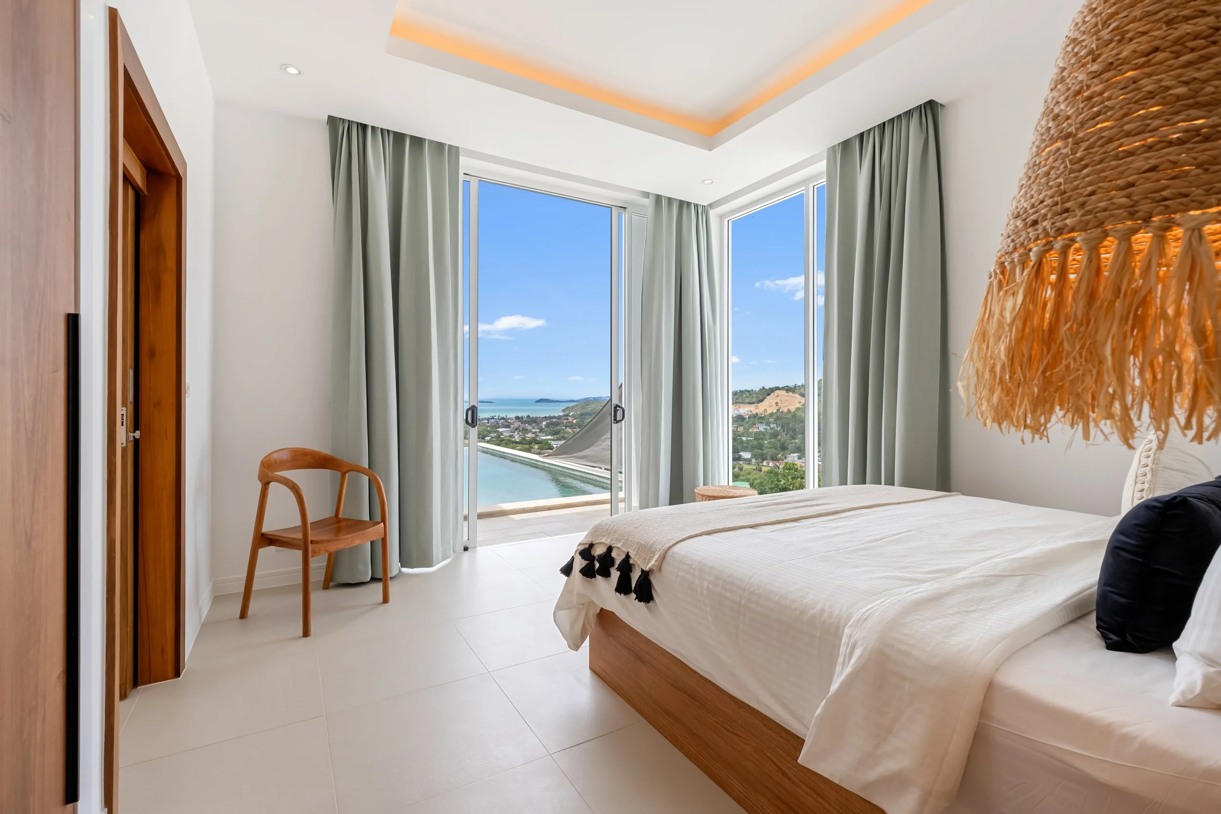 A modern bedroom with large windows overlooking an ocean view, white walls, a beige bed with black and beige accents, a wicker hanging lamp, wooden furniture, and curtains.