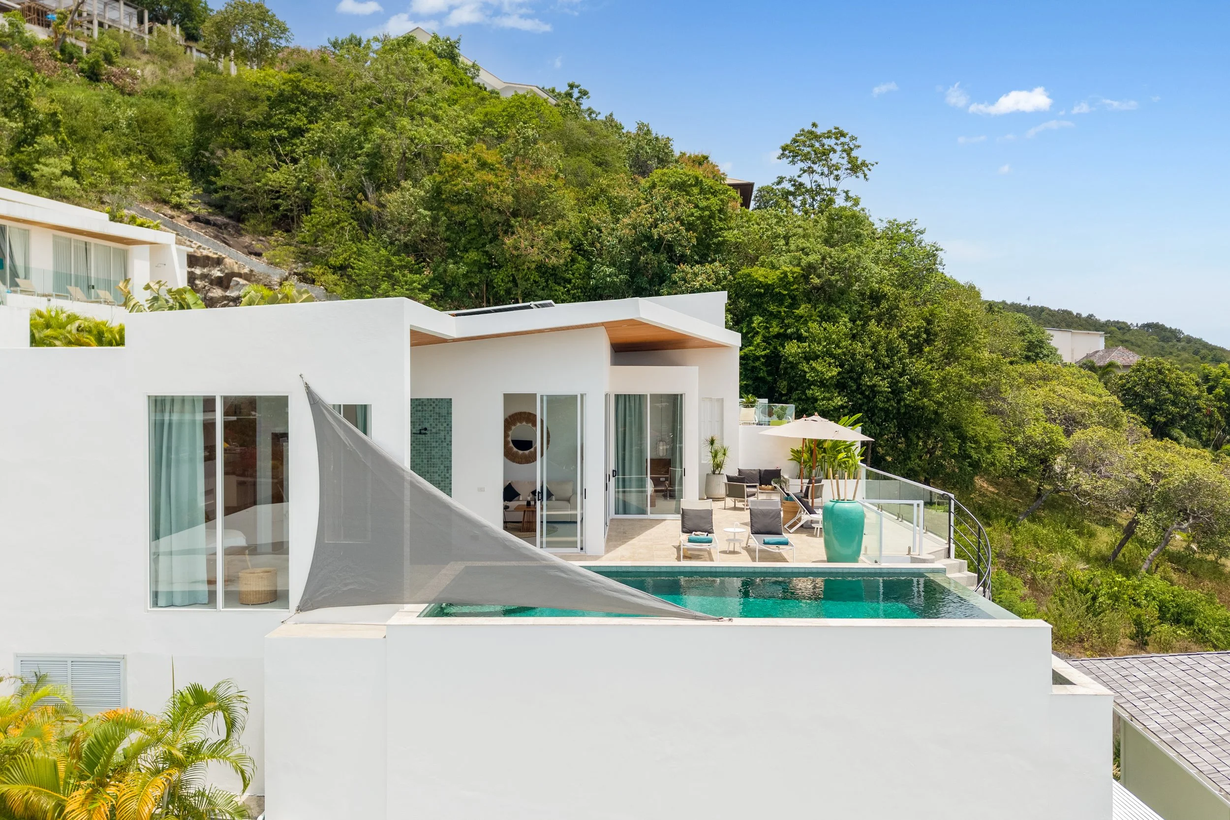 Modern white house on a hillside with outdoor patio, swimming pool, lounge chairs, potted plants, and a canopy, surrounded by lush green trees and a blue sky.
