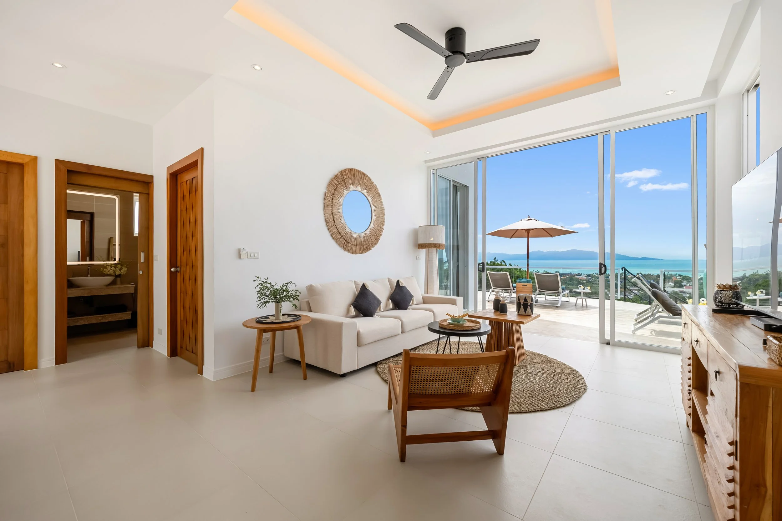 Living room with white sofa, wooden side tables, and a large sliding glass door leading to a balcony with outdoor chairs and an umbrella, overlooking a body of water and distant mountains.