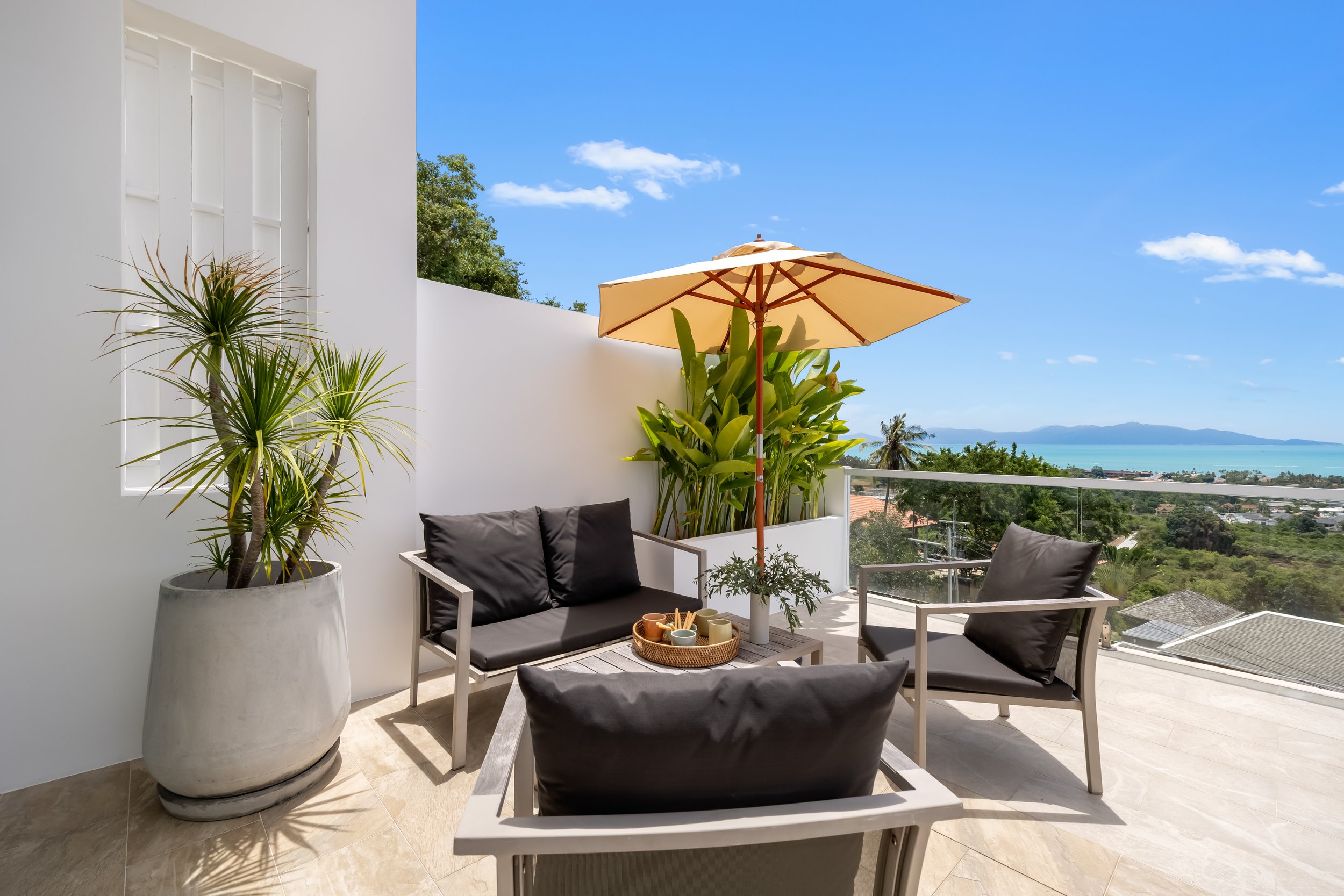 Balcony with white walls and patio furniture, including two chairs with black cushions, a small table with colorful cups, a large potted plant, an umbrella, and a view of a coastal landscape with trees, houses, and blue ocean under a clear sky.