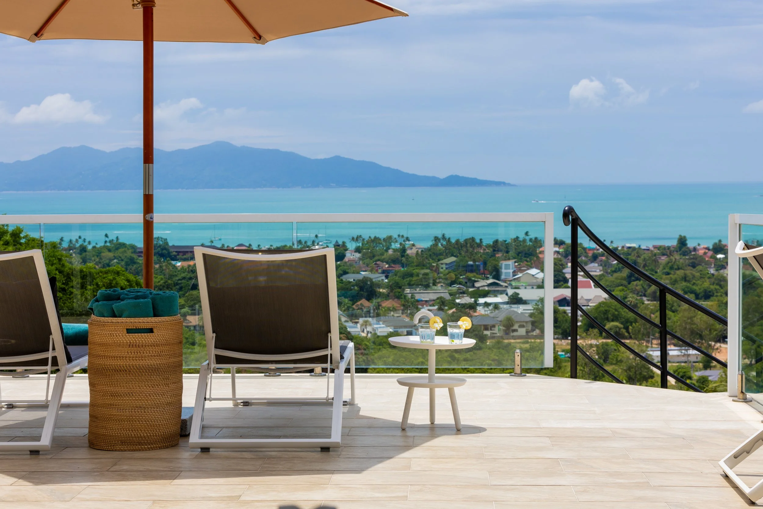 Outdoor balcony with two chairs, a basket with turquoise towels, a small round table with two glasses of water with lemon slices, an opened umbrella, and a scenic view of mountains, ocean, and residential neighborhood under a partly cloudy sky.