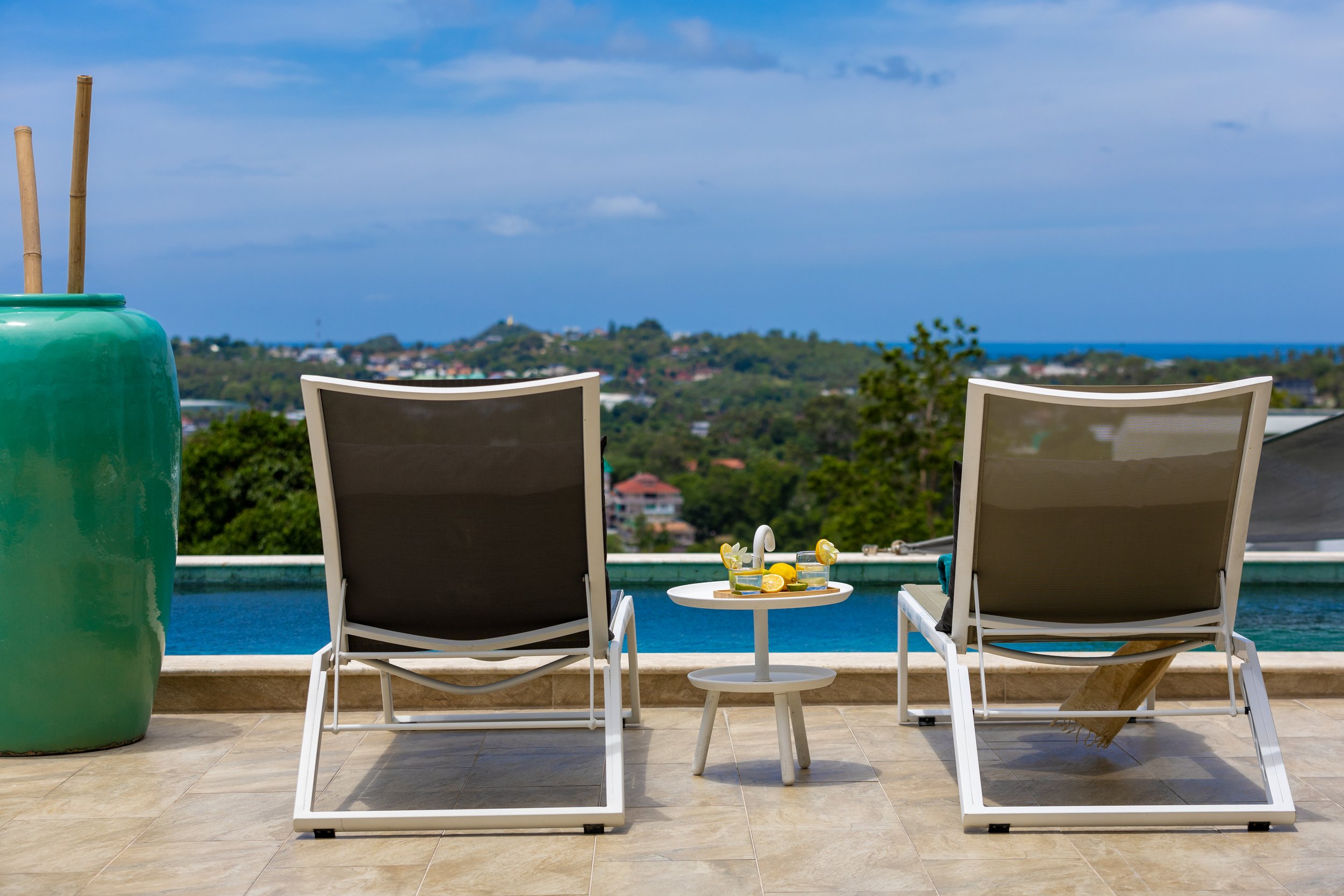 A balcony with two lounge chairs facing a view of a hilly landscape and the ocean in the distance. There are two glasses of lemonade and lemons on a small white table between the chairs, and a large green ceramic vase with wooden sticks on the left.