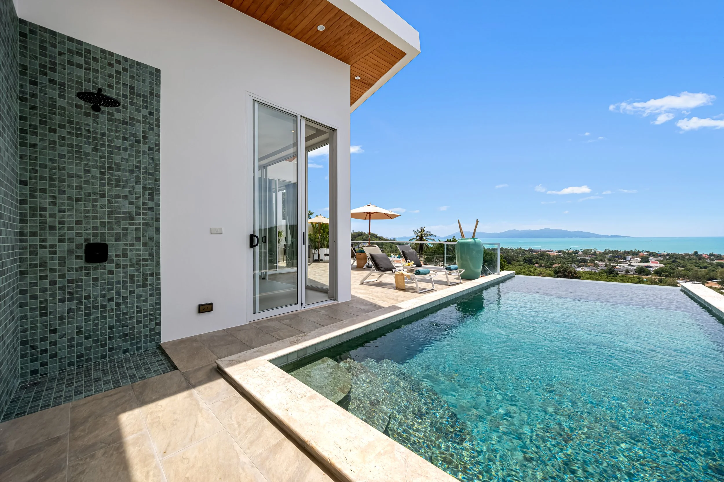 Luxury house balcony with swimming pool, lounge chairs, umbrellas, and ocean view in the background.