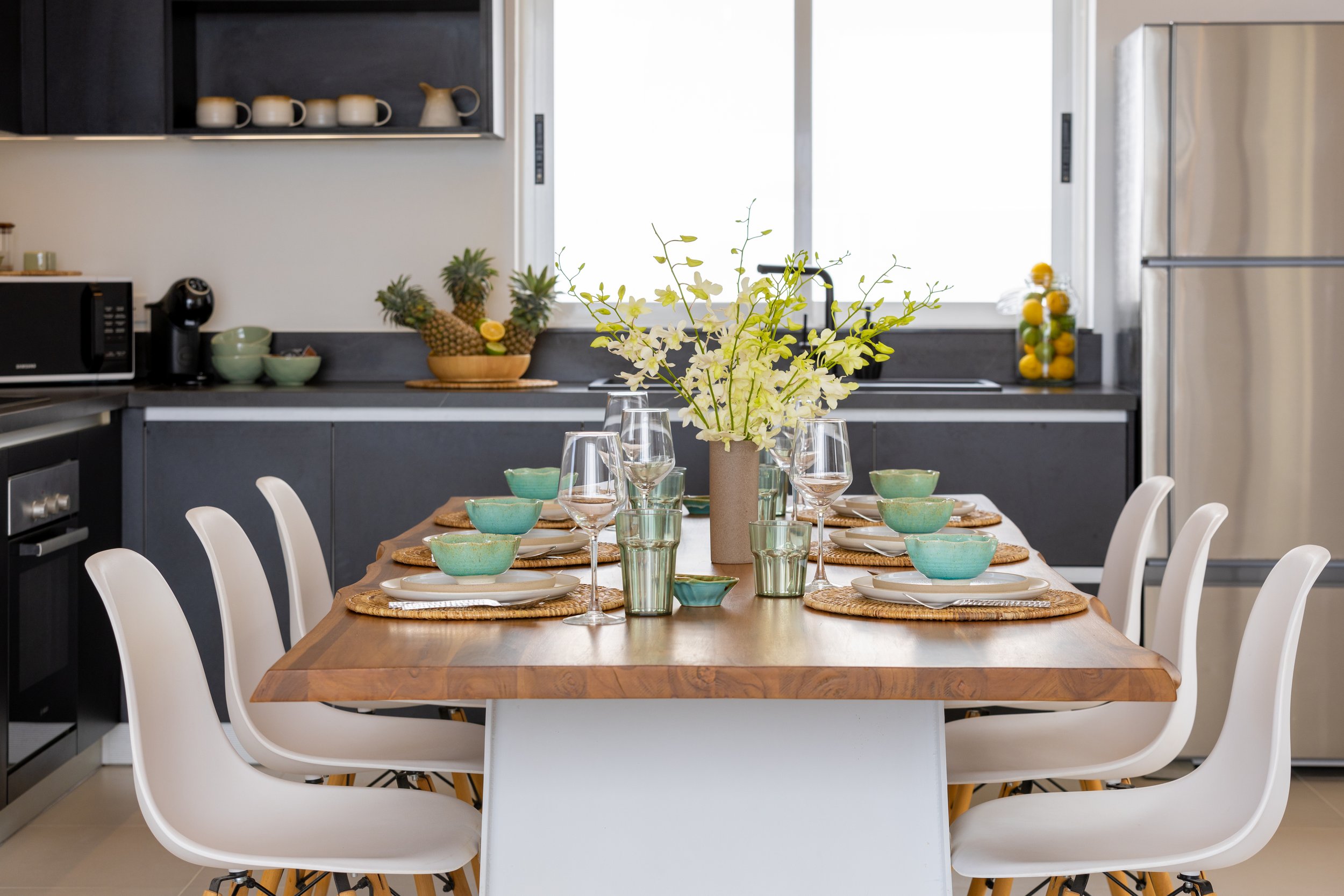 A dining table set with turquoise bowls, glasses, and woven placemats in a modern kitchen with black cabinets and a large window.