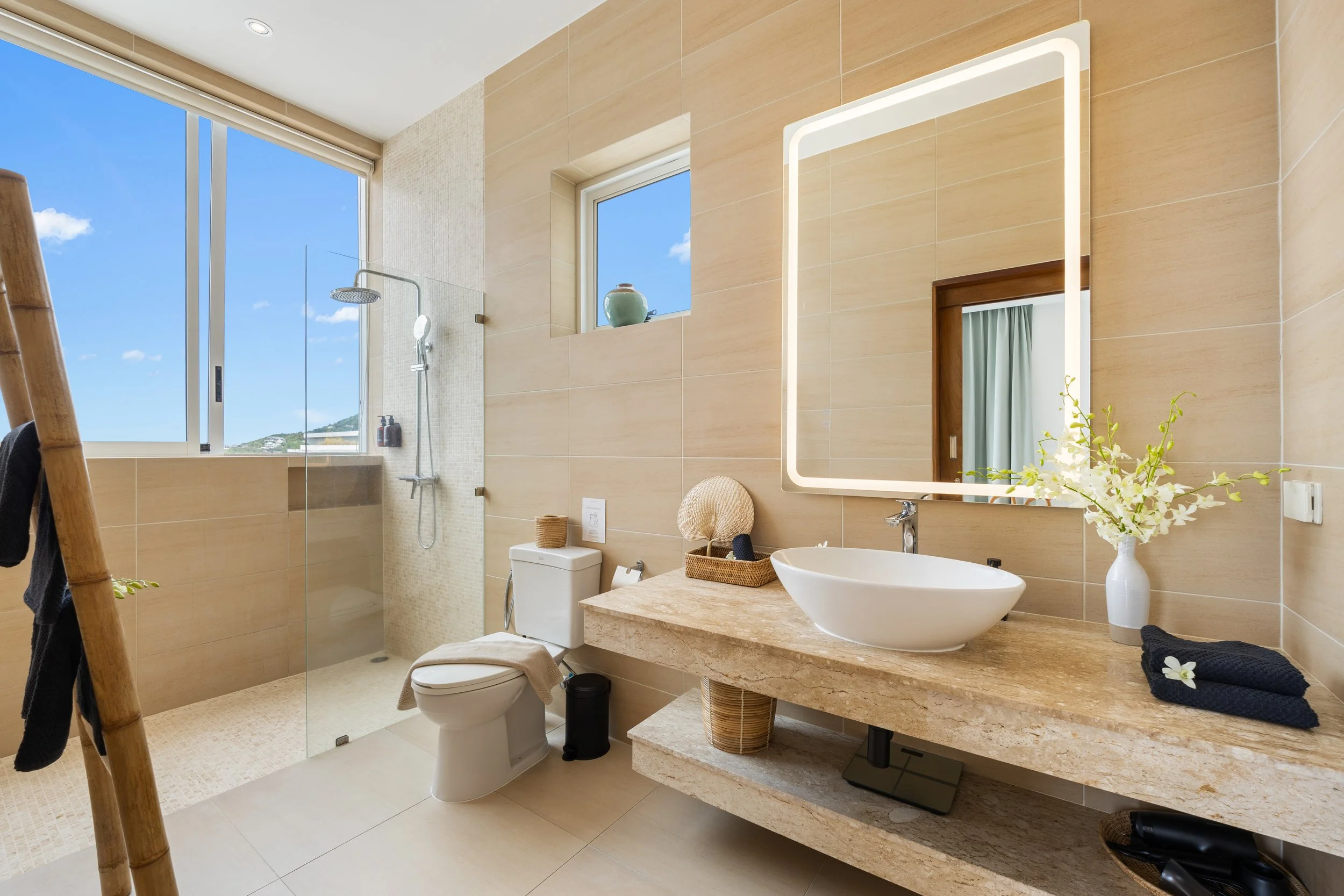 Modern bathroom with large window, beige tile walls, and a wooden vanity with a white vessel sink and a large mirror with LED lighting. Glass shower enclosure with rainfall showerhead, toilet, and decorative flowers in a vase.