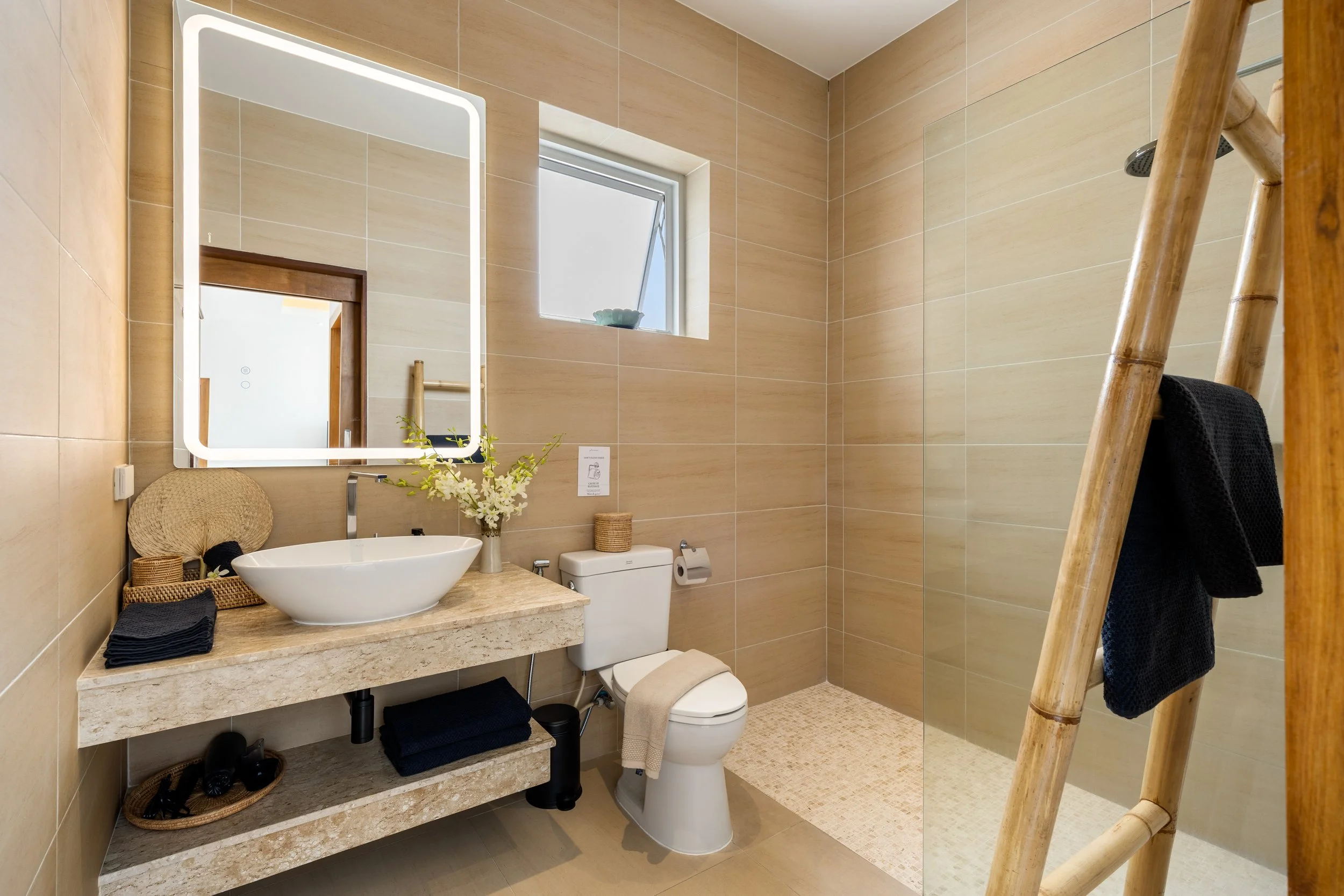 Modern bathroom with beige tiled walls and floor, a mirror with built-in lighting above a stone countertop sink, a small window, a toilet, and a wooden ladder with a black towel hanging on it.