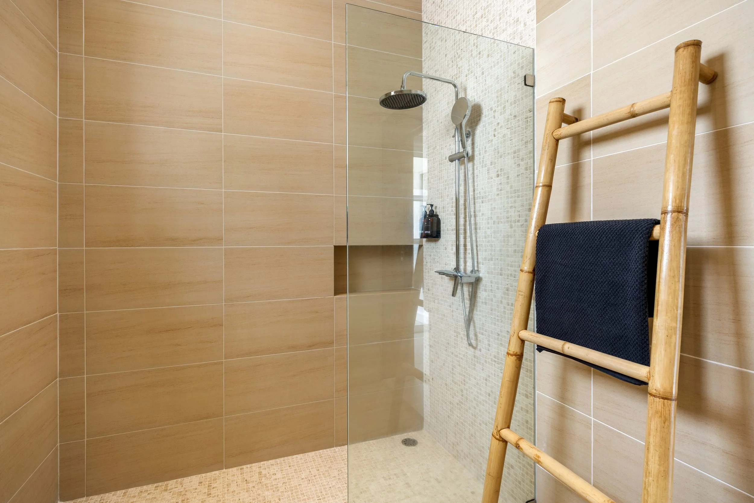 A modern walk-in shower with beige large tile walls, a glass partition, a rain shower head, a handheld shower head, and a wooden towel rack with a black towel.