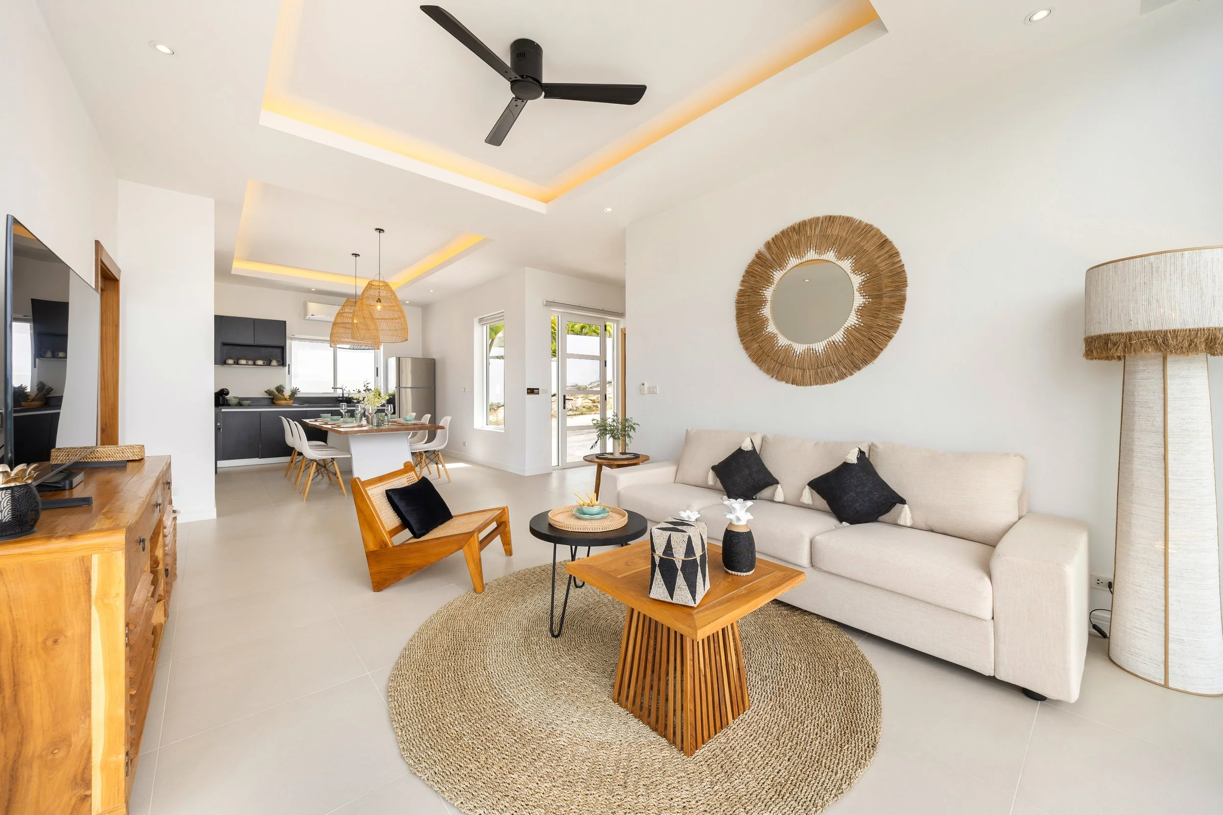 A modern, open-concept living room with a beige sofa, black and white pillows, wooden side tables, a woven rug, and tropical decor. The room features a high ceiling with recessed lighting, a ceiling fan, a large round mirror with a woven frame, and a floor lamp with a woven shade. In the background, an adjacent kitchen and dining area have dark cabinets, a window, and a round wooden dining table with chairs.