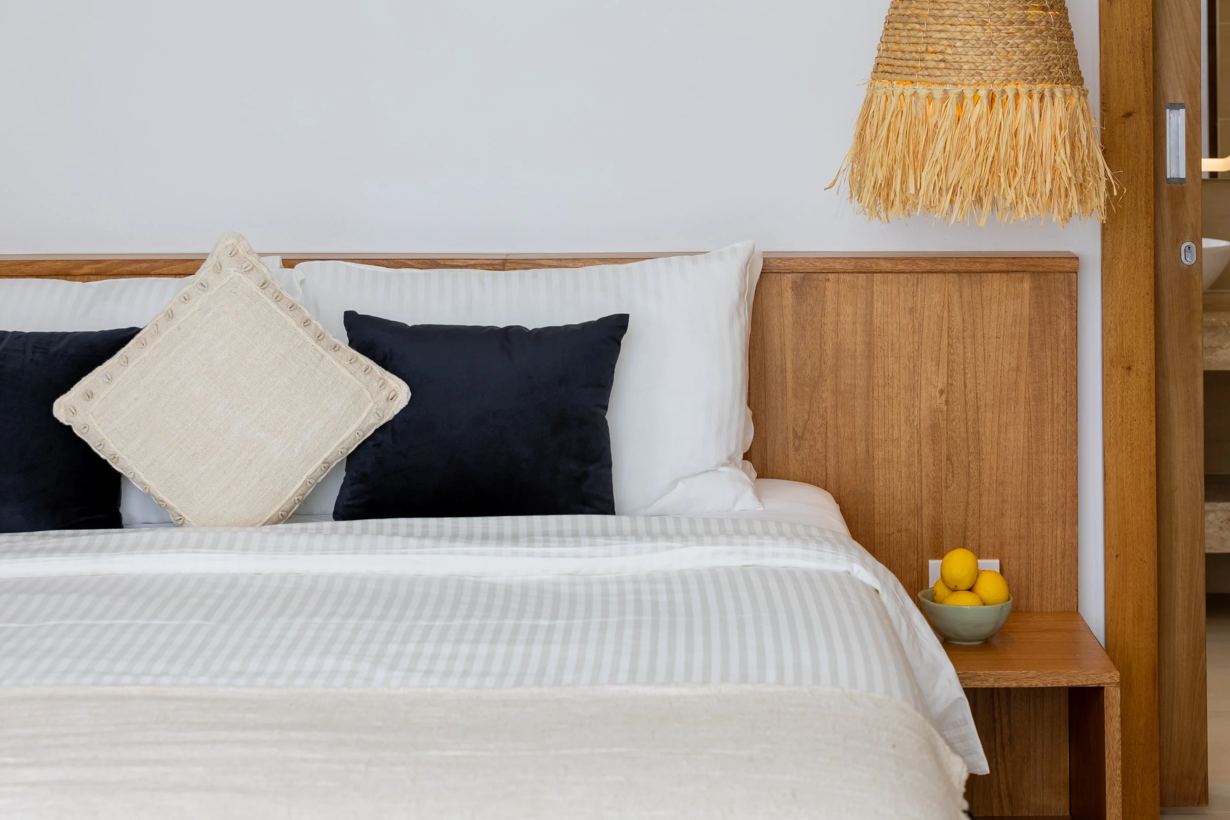 Close-up of a bed with white bedding, four pillows of different colors and textures (white, beige, black), and a wicker lampshade hanging above a wooden headboard with a built-in bookshelf.