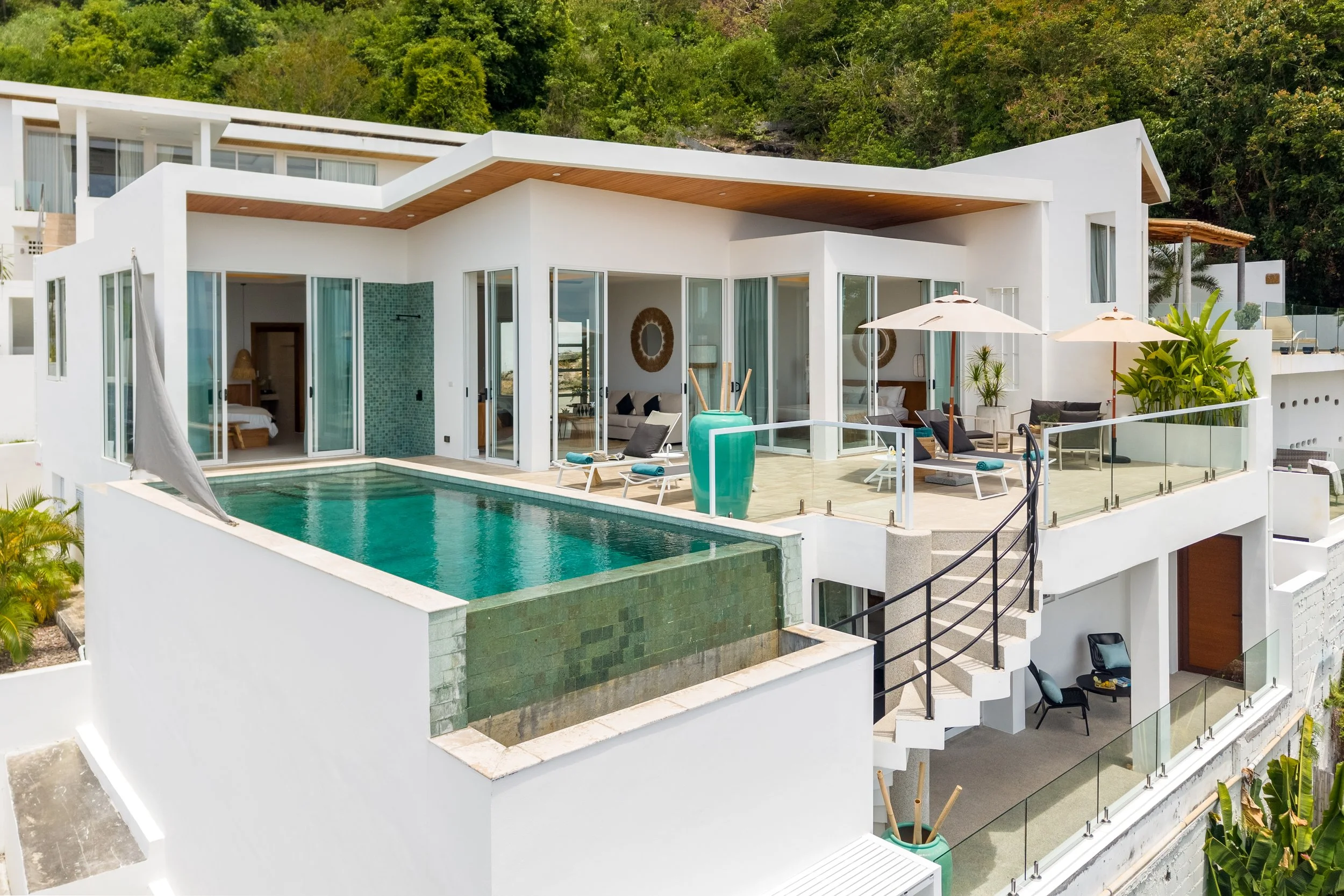 Modern white multi-story house with large glass windows and sliding doors, outdoor pool, patio furniture, umbrellas, and lush green hillside in the background.