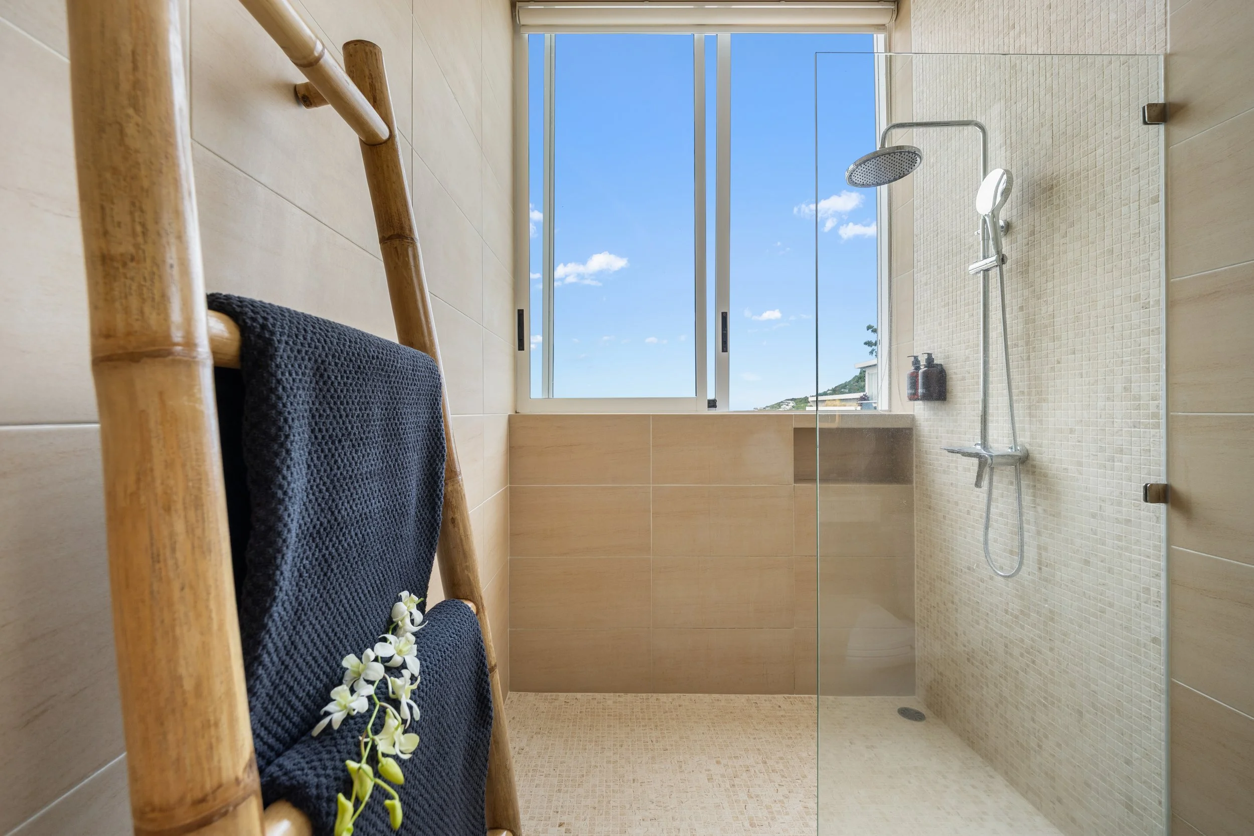 Interior view of a modern bathroom with a walk-in shower, a large window, a wooden towel rack with a dark towel and white flowers, and a blue sky outside.