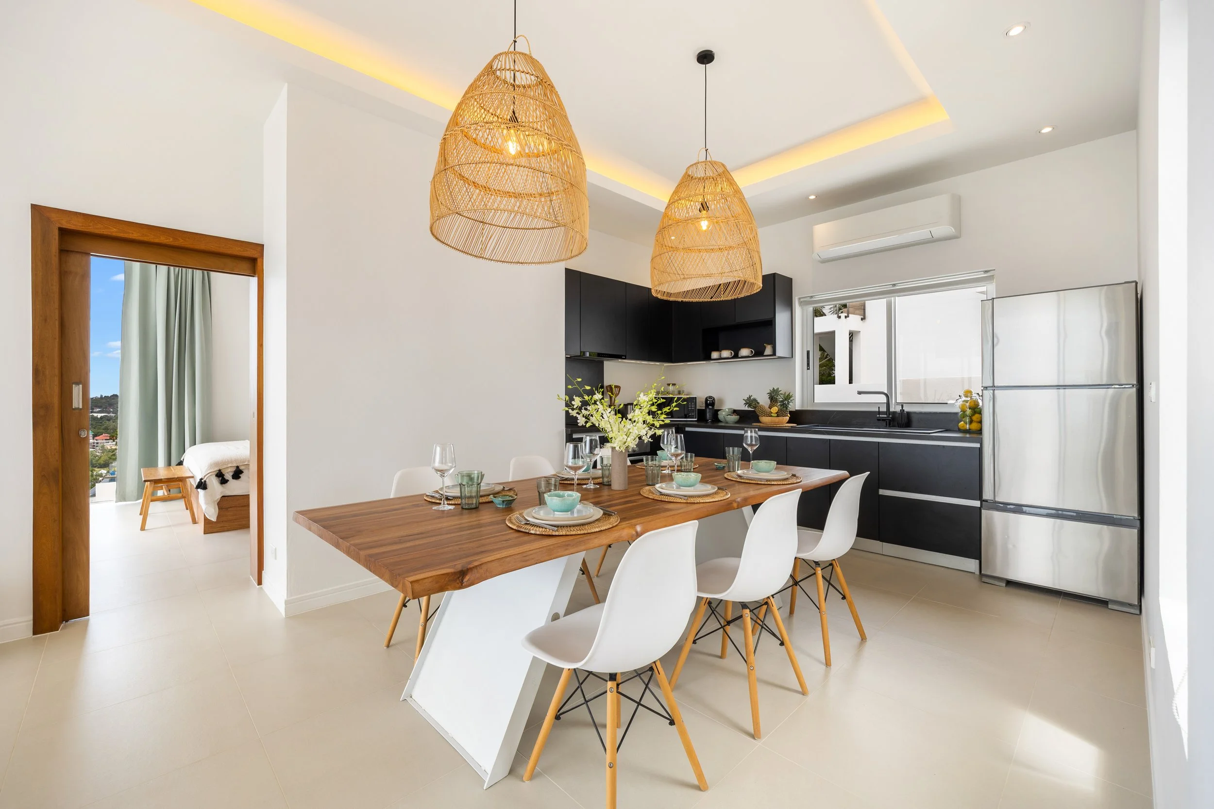 Modern kitchen and dining area with white walls, wood and black cabinetry, a wooden dining table set with plates and glasses, white chairs, wicker pendant lights, and large windows with outdoor views.