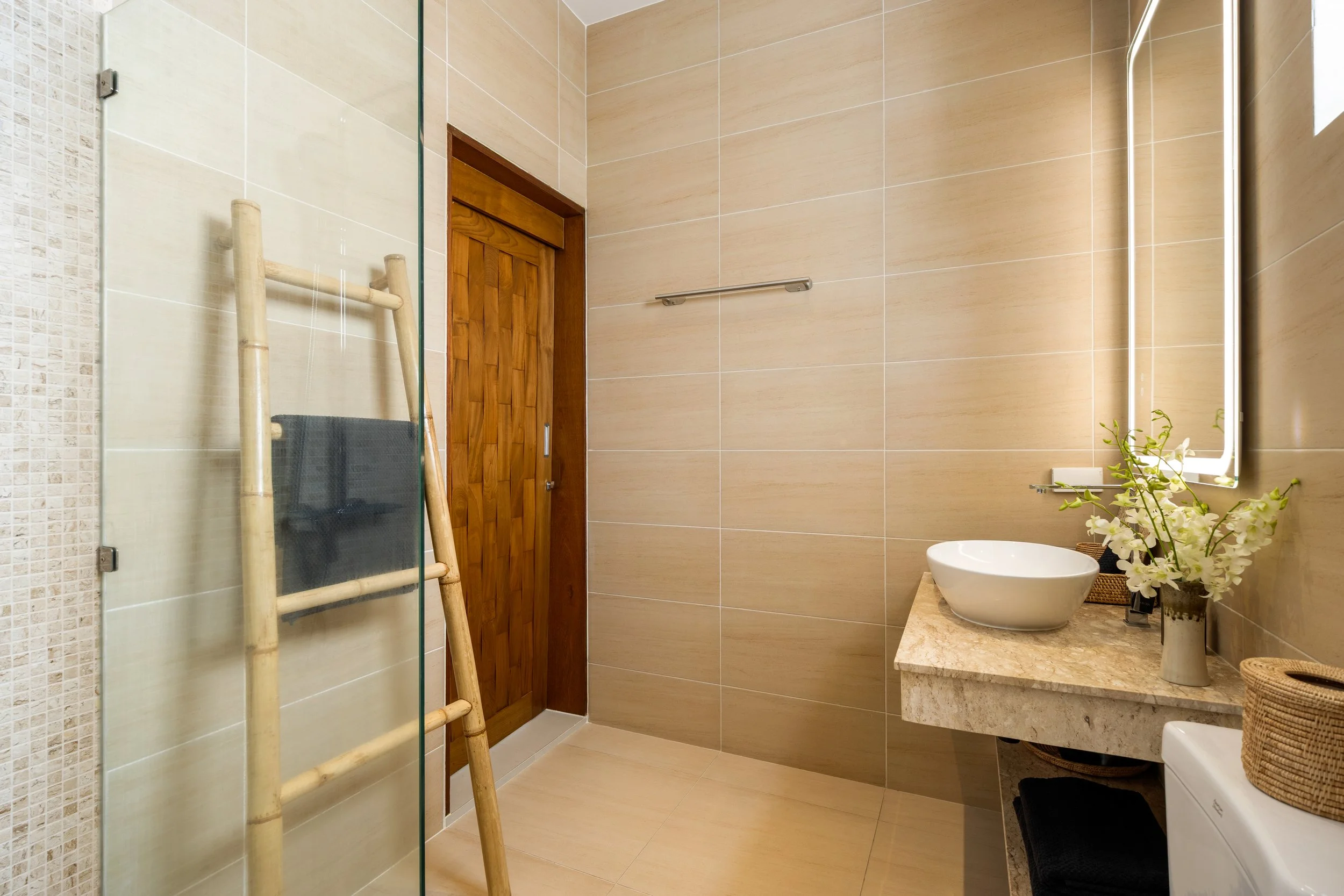 A modern bathroom with beige tiled walls and a mirror above a beige marble countertop. On the countertop, there is a white vessel sink and a plant with white flowers. A wooden door is visible, along with a bamboo ladder and towel, and a washing machine underneath the counter.