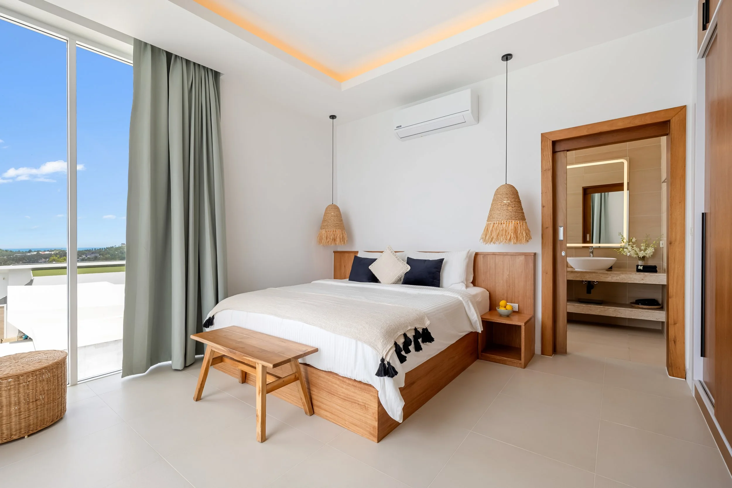 Modern hotel room with a large bed, wooden bed frame, and minimal decor. Large glass sliding door with green curtains showing a view of blue sky and greenery outside. Two woven pendant lights hang above the bed, and a small wooden bench is at the foot of the bed. A bathroom with a mirror and sink is visible through a wooden frame opening.