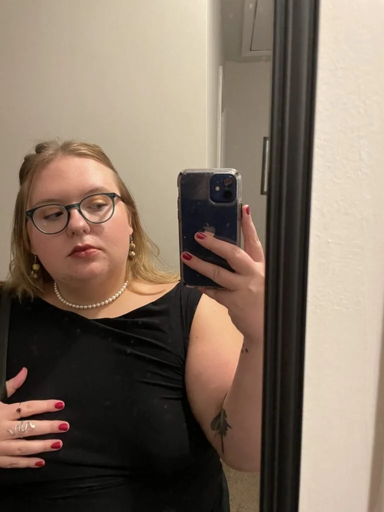 A woman with glasses, pearl necklace, and earrings takes a mirror selfie wearing a black sleeveless top. She has blonde hair and red painted nails.