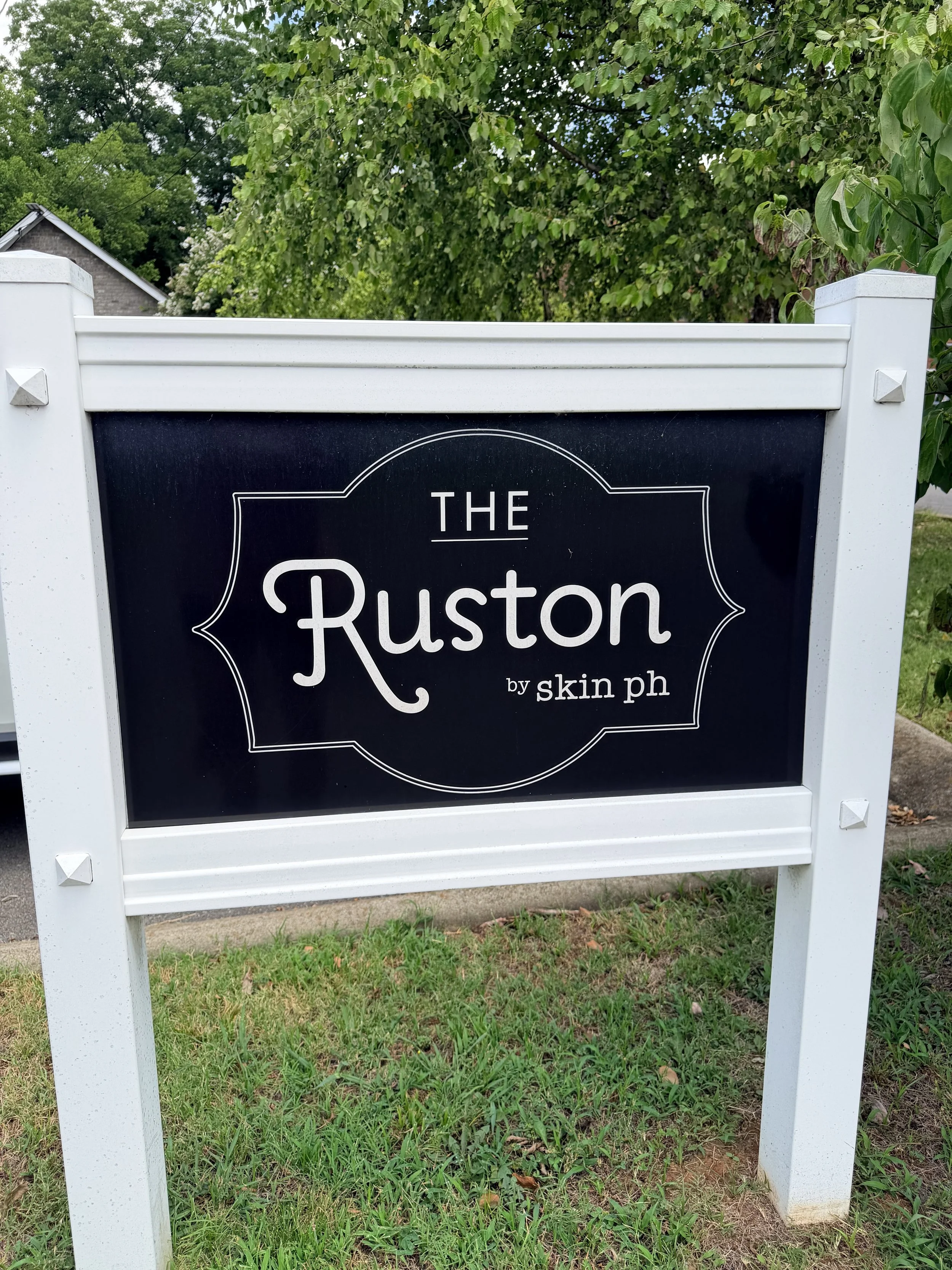 A black and white outdoor signboard on a white frame with the text 'THE Ruston by skin ph,' situated on grass with trees and a house in the background.
