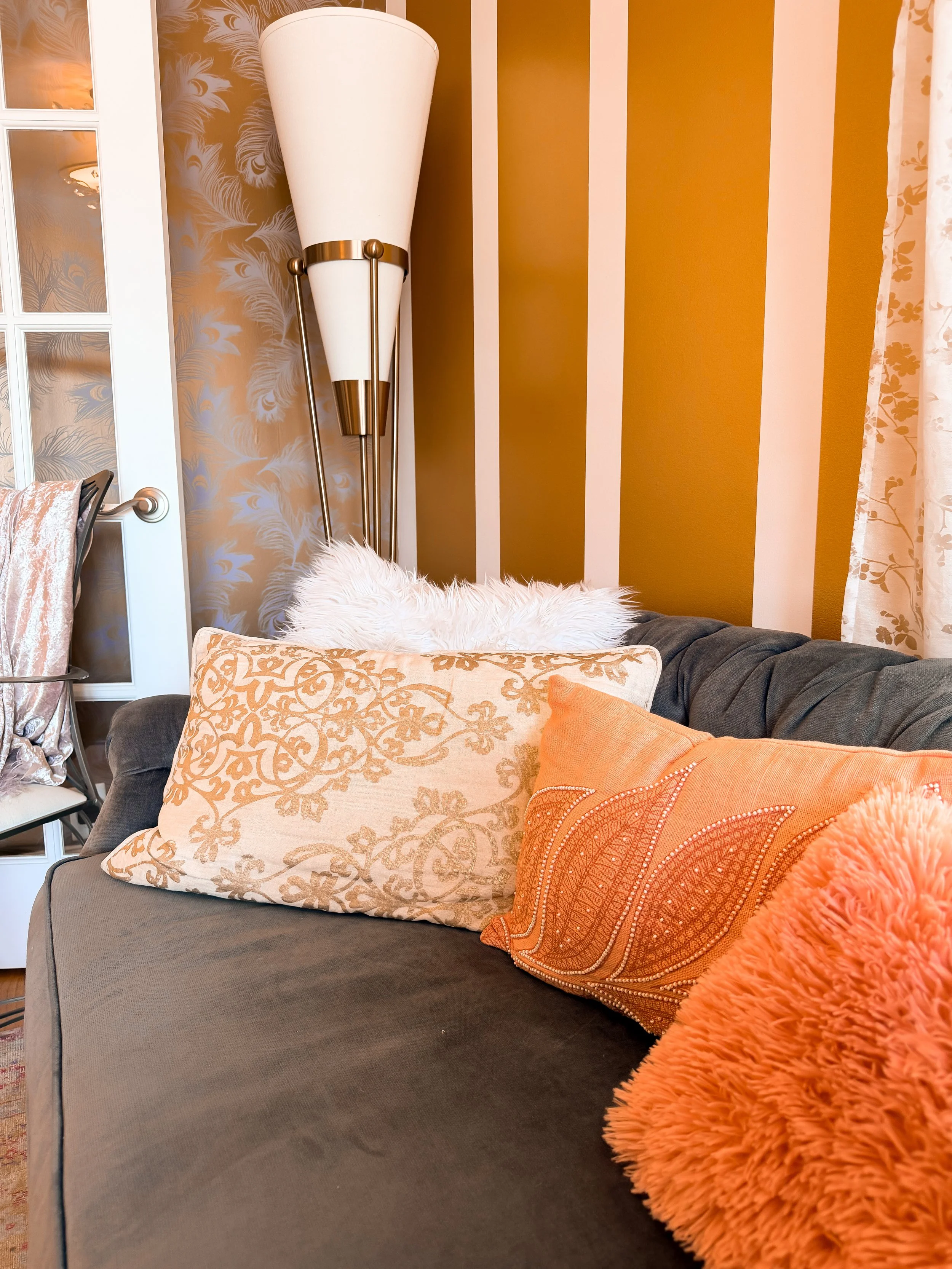 A cozy living room corner with a grey sofa, decorative pillows in orange and beige patterns, a fluffy orange throw blanket, a white table lamp with gold accents, a floral curtain on the right, and a wall with gold and beige vertical stripes and leaf-patterned wallpaper.