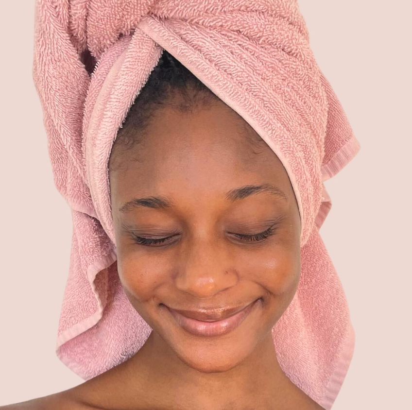 A woman with a pink towel wrapped around her head, smiling with her eyes closed, skin appears clean and radiant.