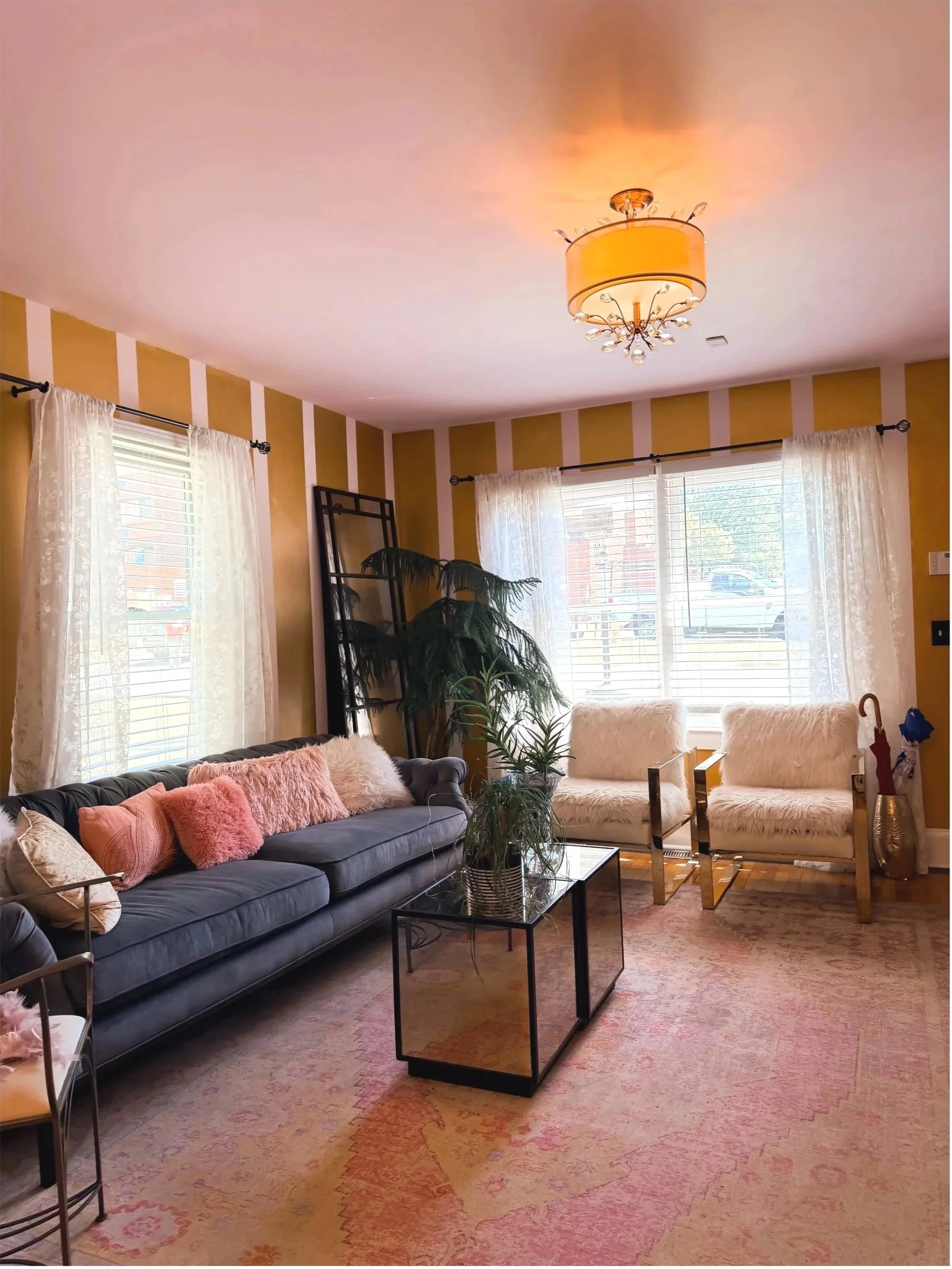 Living room with beige and white striped walls, large windows with sheer curtains, a dark blue sofa with pink and white pillows, white armchairs with fluffy cushions, a glass-top coffee table with a potted plant, a pink-patterned area rug, a ceiling light fixture with a drum shade and decorative crystals, and some houseplants