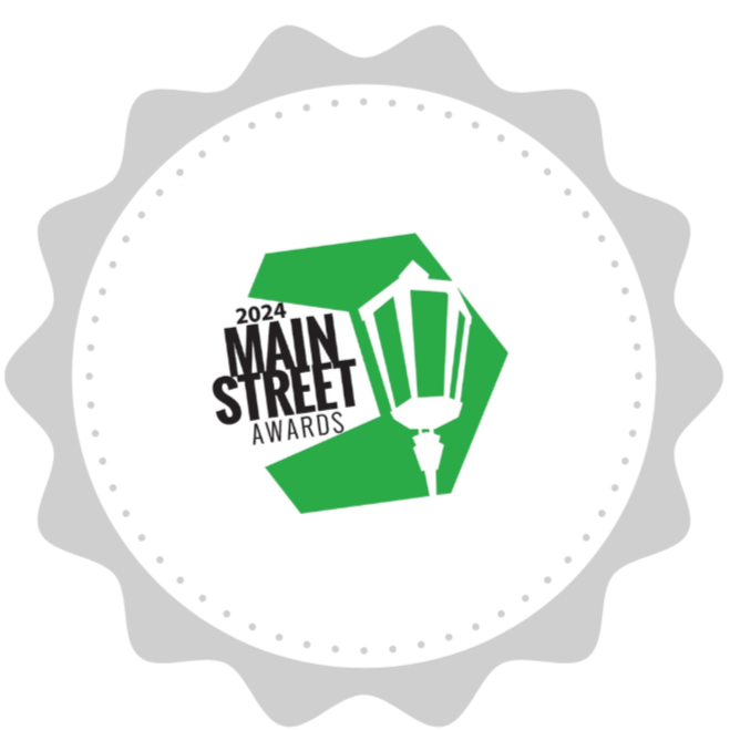 A circular badge with a scalloped edge featuring a green street lamp graphic and text that reads "2024 Main Street Awards."