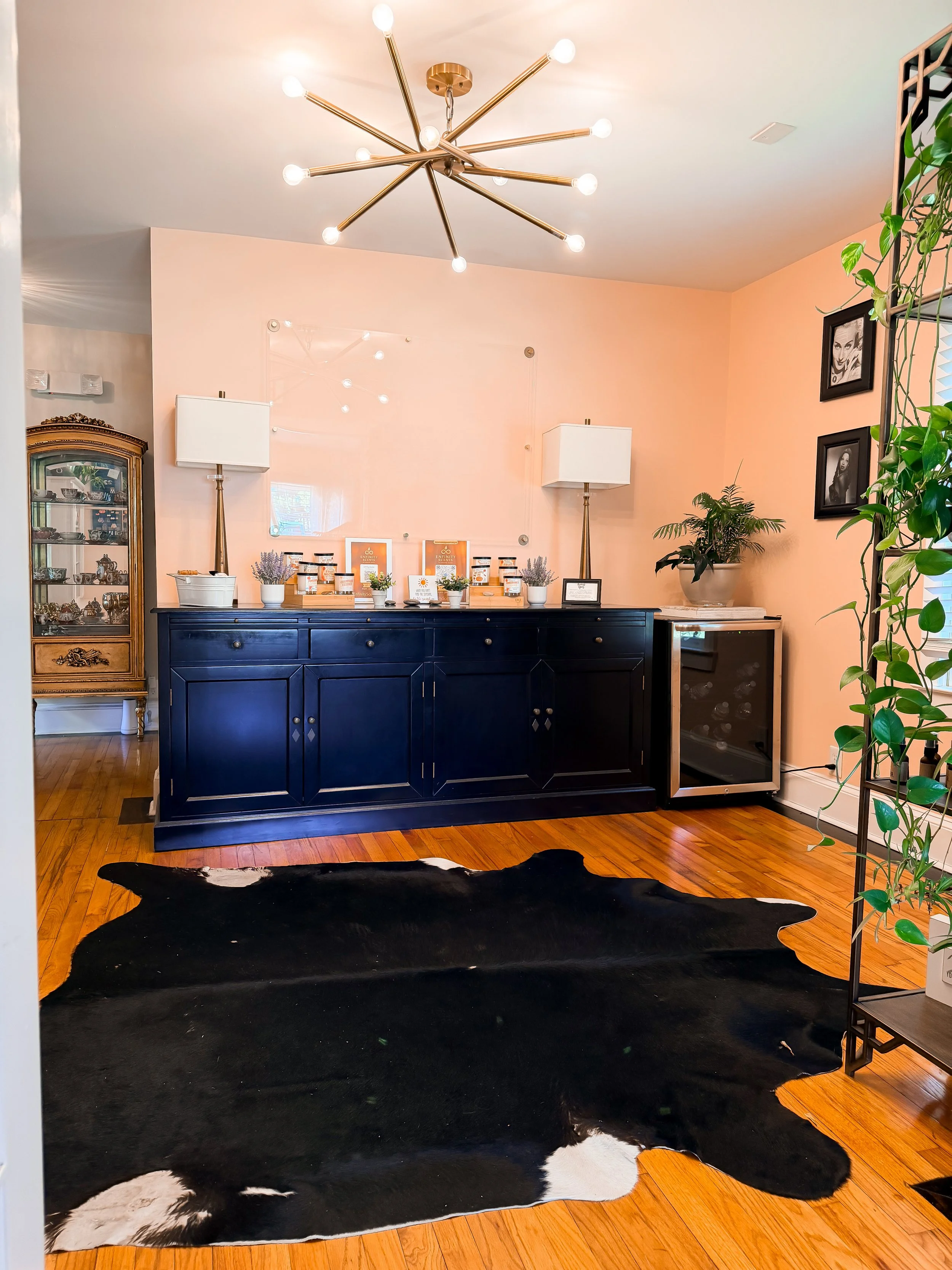 Living room with a black cowhide rug on wooden floor, a dark blue sideboard with decorative items and framed pictures on wall, plants, and modern lighting fixtures with exposed bulbs.