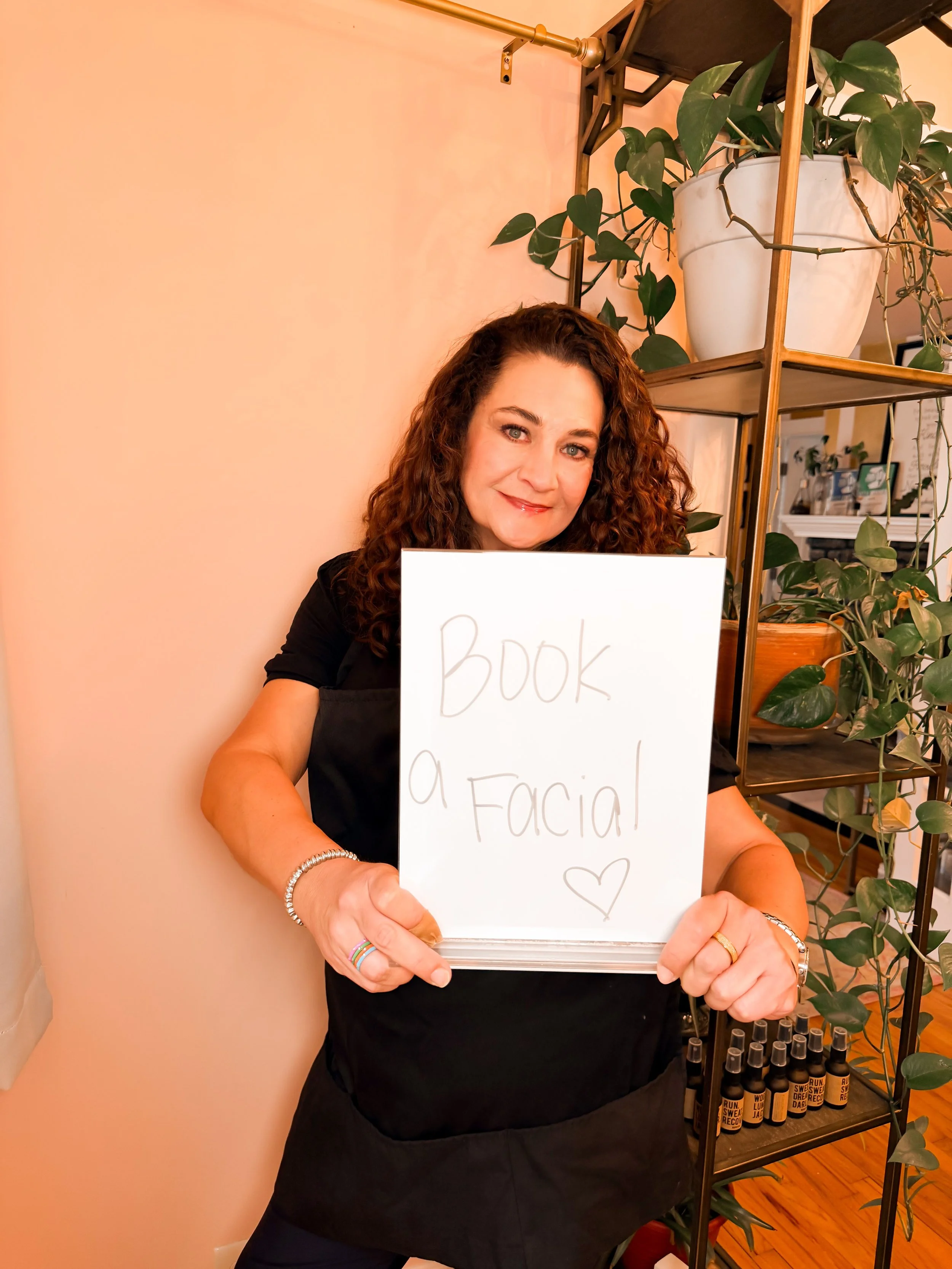 A woman with curly brown hair smiling while holding a whiteboard with 'Book a Facial' written on it. She is standing indoors near a plant and a wooden shelf with bottles underneath.