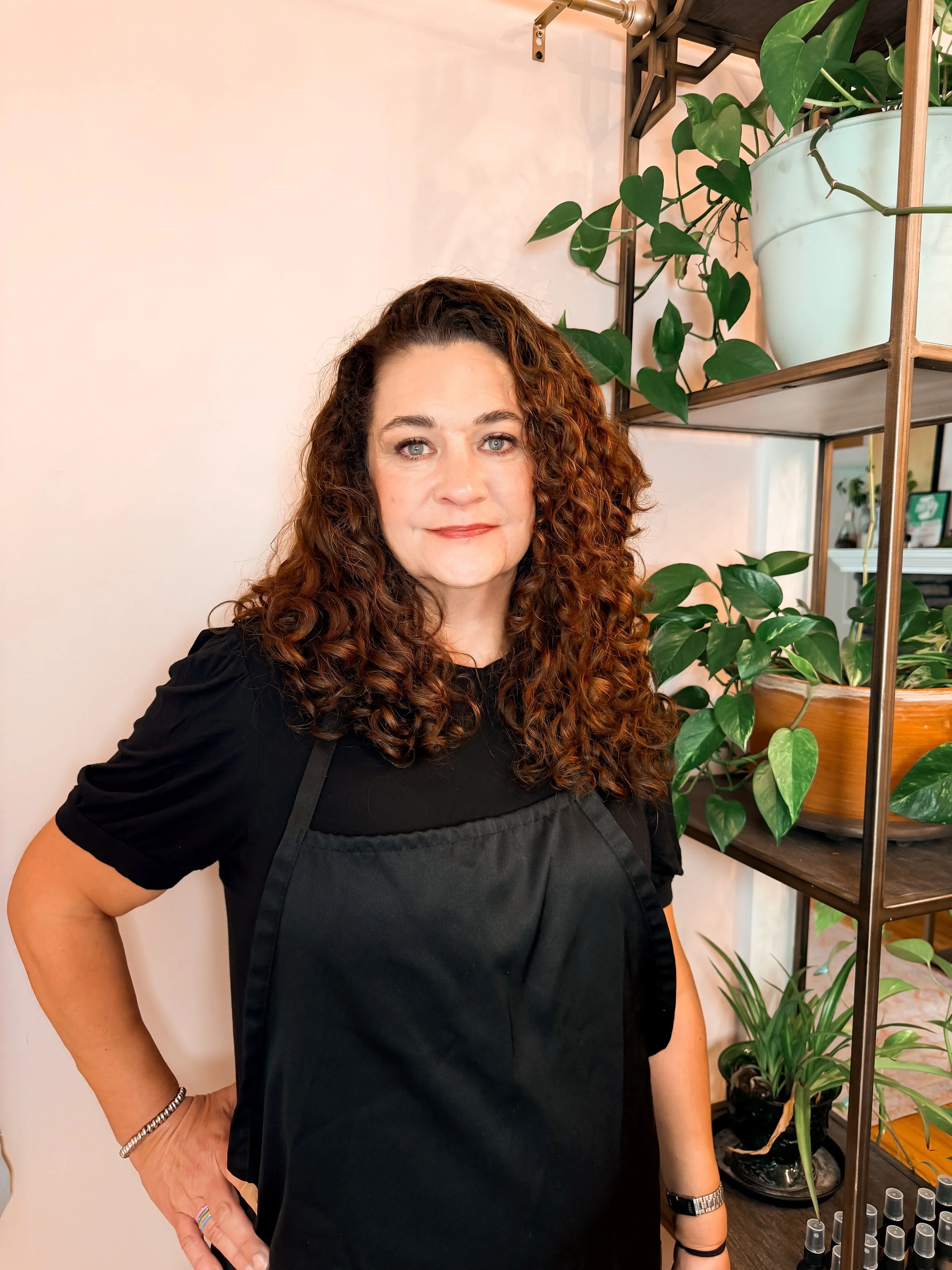 Keisha has curly brown hair and light skin standing next to a plant shelf with green plants in pots, wearing a black top, silver bracelet, and watch, in a room with a light-colored wall.