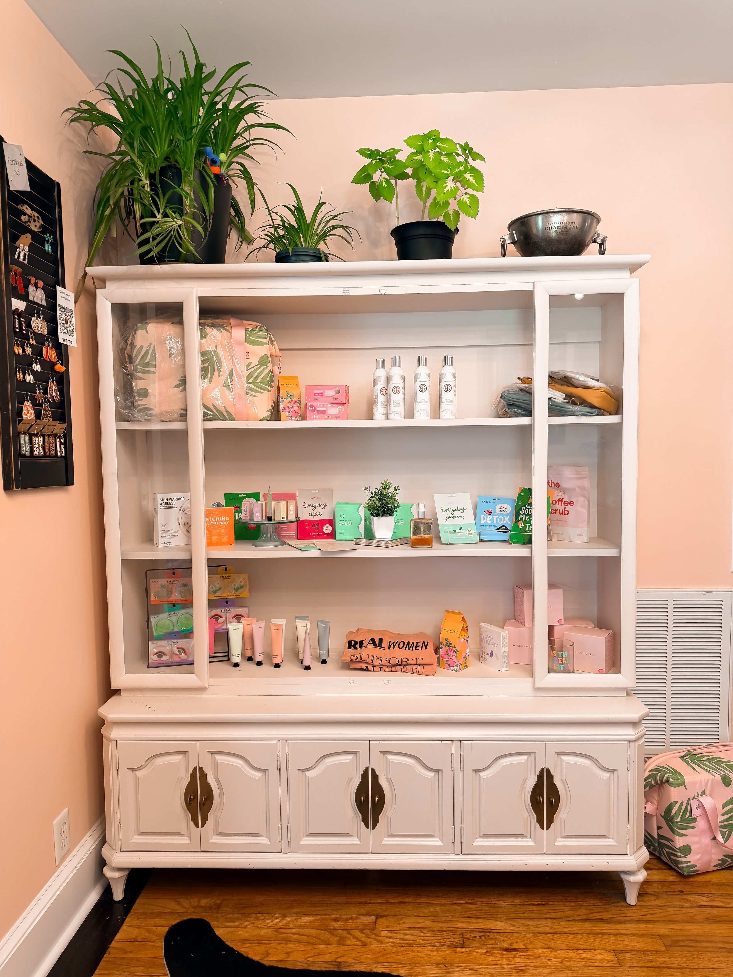 White vintage cabinet with glass doors displaying skincare products, supplements, and decorative items in a room with pink walls and hardwood floors.