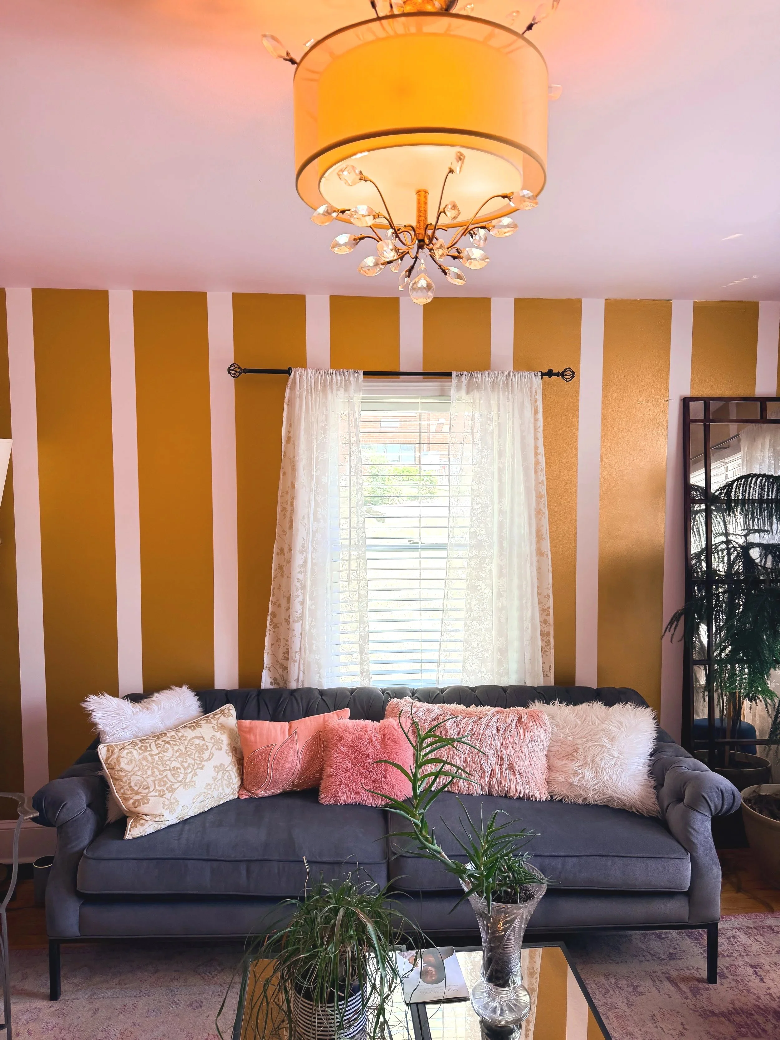Living room with a dark gray sofa decorated with pink, beige, and furry pillows. A glass coffee table in front of the sofa holds a small potted plant and a book. Behind the sofa is a window with white curtains, and the wall features bold vertical stripes in yellow and white. A yellow chandelier with crystal accents hangs from the ceiling, and part of a tall plant is visible on the right side of the image.