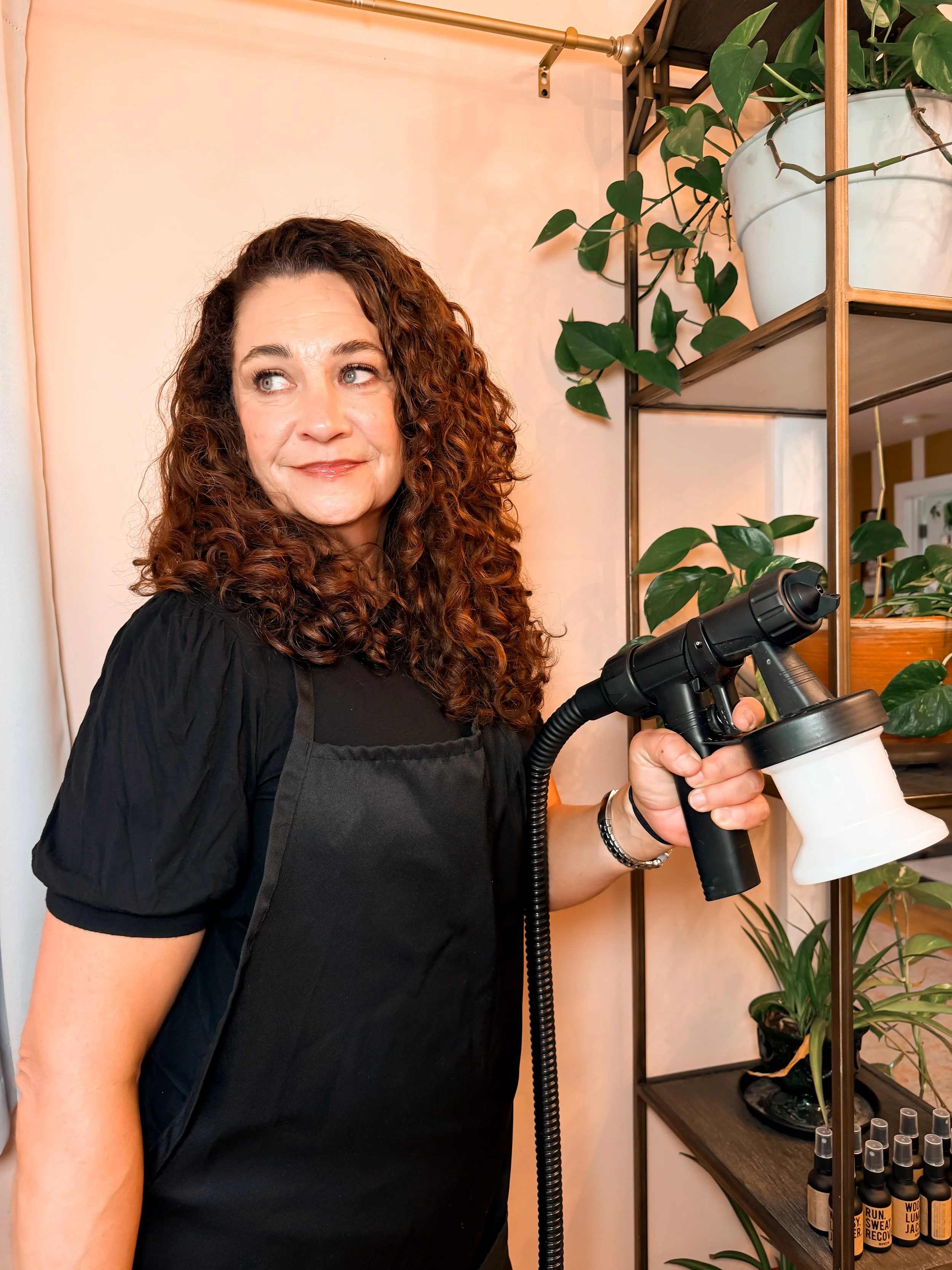 A woman with curly hair wearing a black shirt holding a disinfectant spray bottle, standing next to indoor plants on a shelving unit.