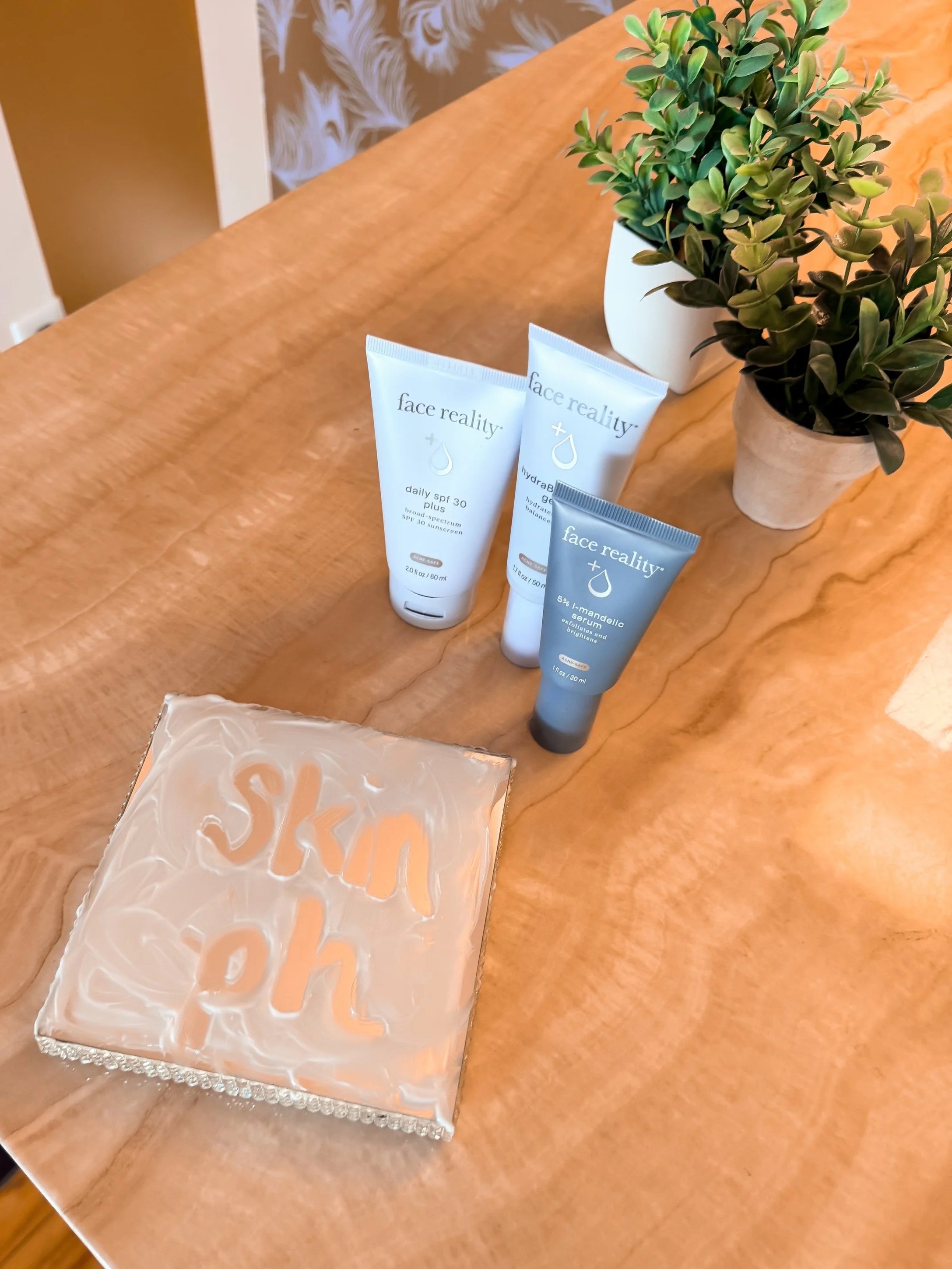 A wooden table with two potted plants and three tubes of skincare products. The products are from the brand face reality, including a sunscreen and serum, with one tube having a blue packaging and the other two in white packaging with gray caps.