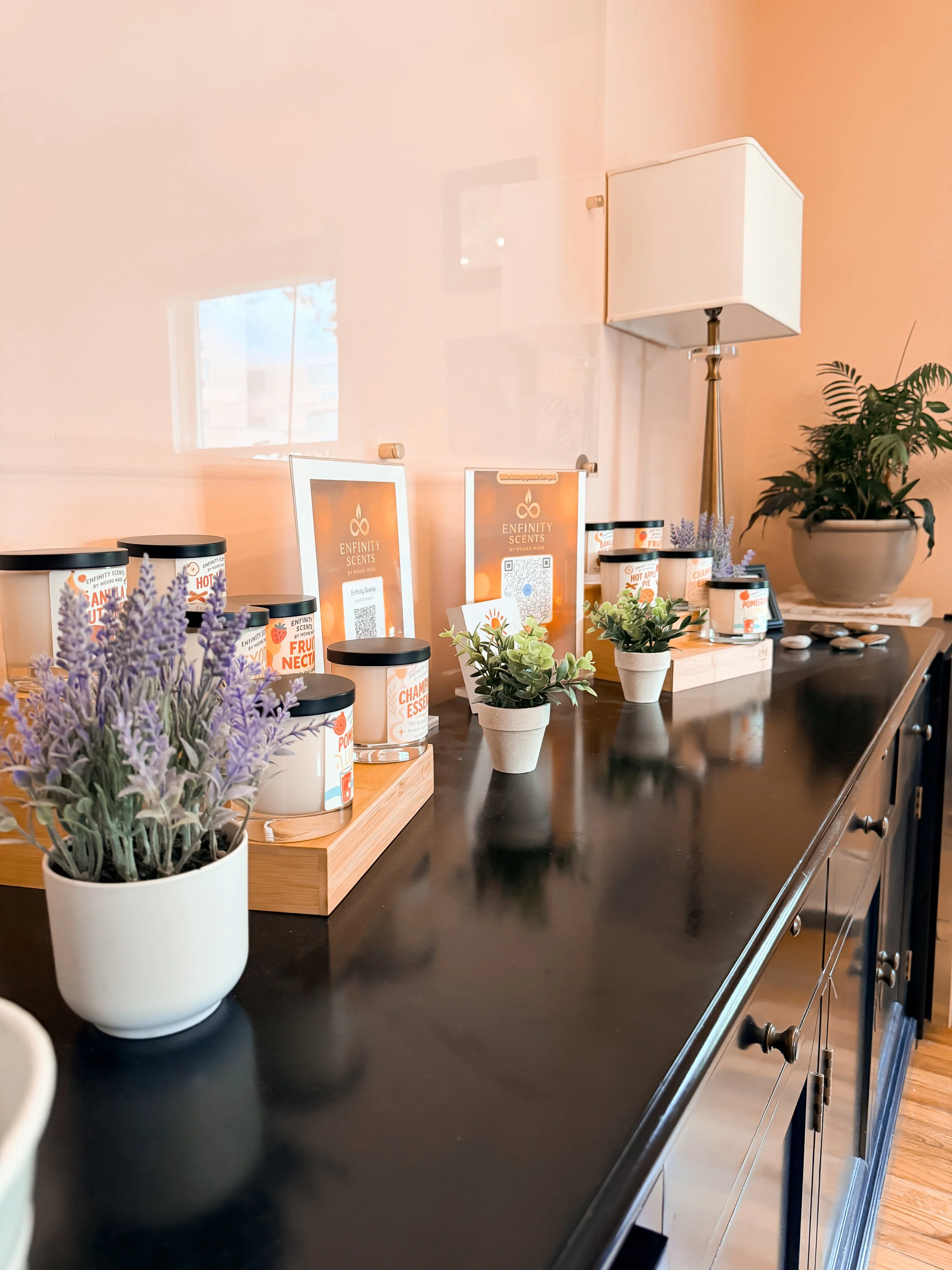 A black countertop with potted lavender and green plants, jars of scented products, and framed informational cards for Enfinity Scents, next to a white table lamp with a rectangular shade and metallic stand, against a peach-colored wall.