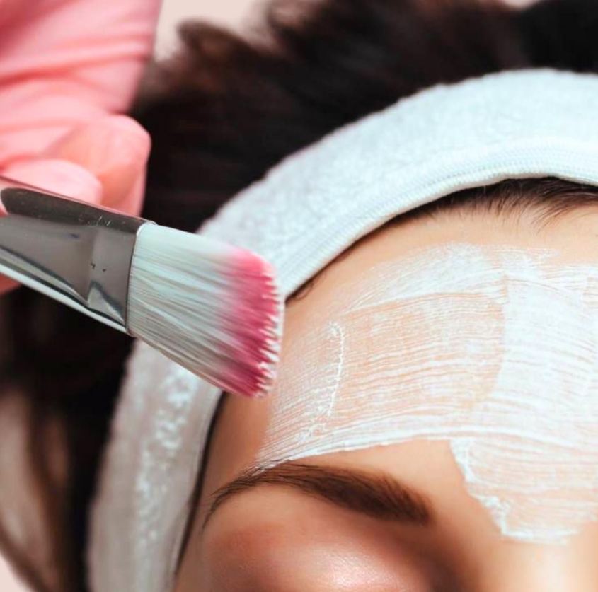 A person applying white facial mask with a paintbrush on their forehead, wearing a headband to hold back hair.