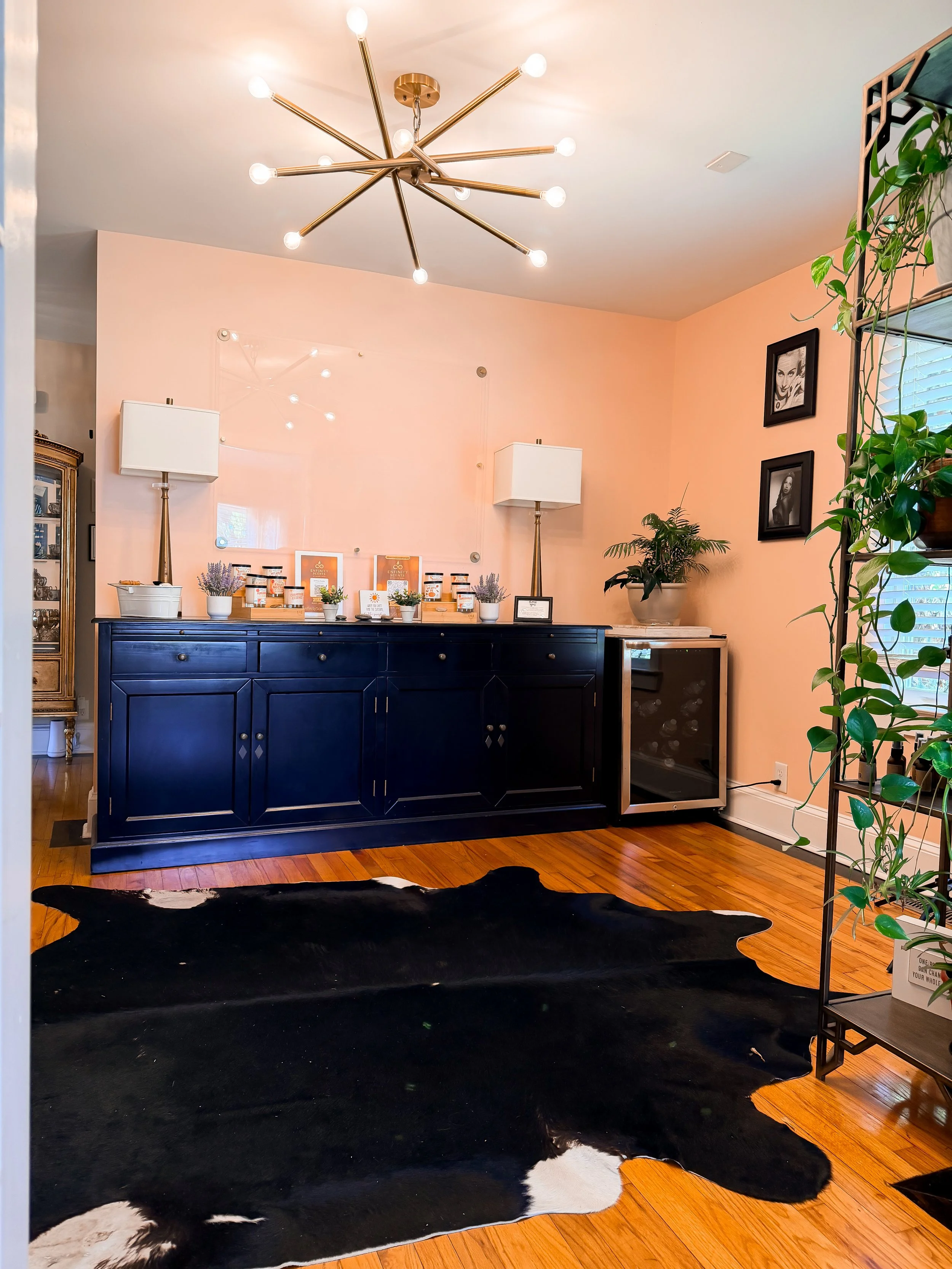 Interior living space with a pink wall, black and white cowhide rug, dark blue sideboard, and plants. Beautiful modern chandelier with multiple arms and light bulbs hangs from white ceiling. Two framed pictures on wall, a large plant in a pot, and a 